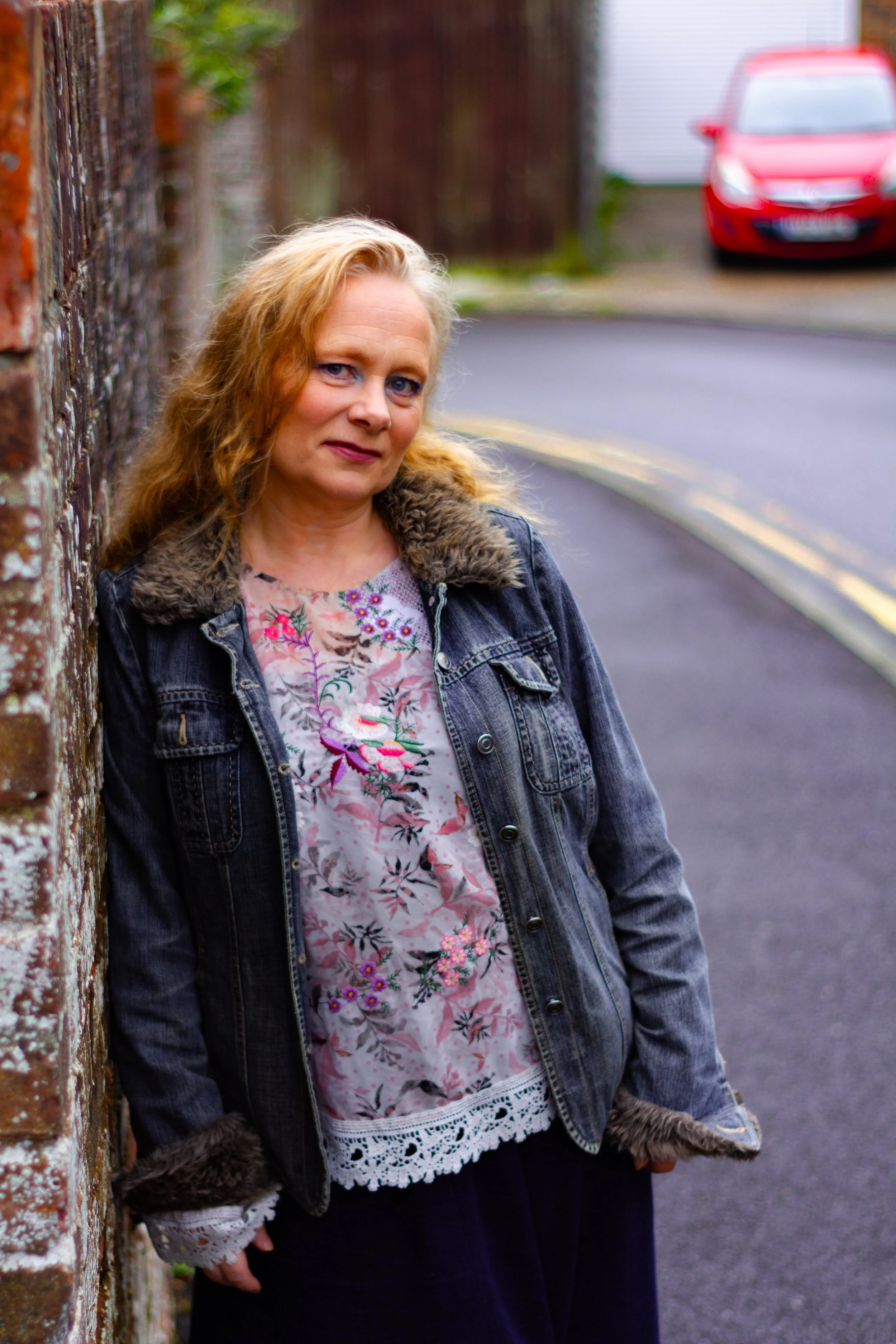 A woman with blonde curly hair leaning against a brick wall on a street. She is wearing a denim jacket with fur trim and a floral blouse. A red car is visible in the background.