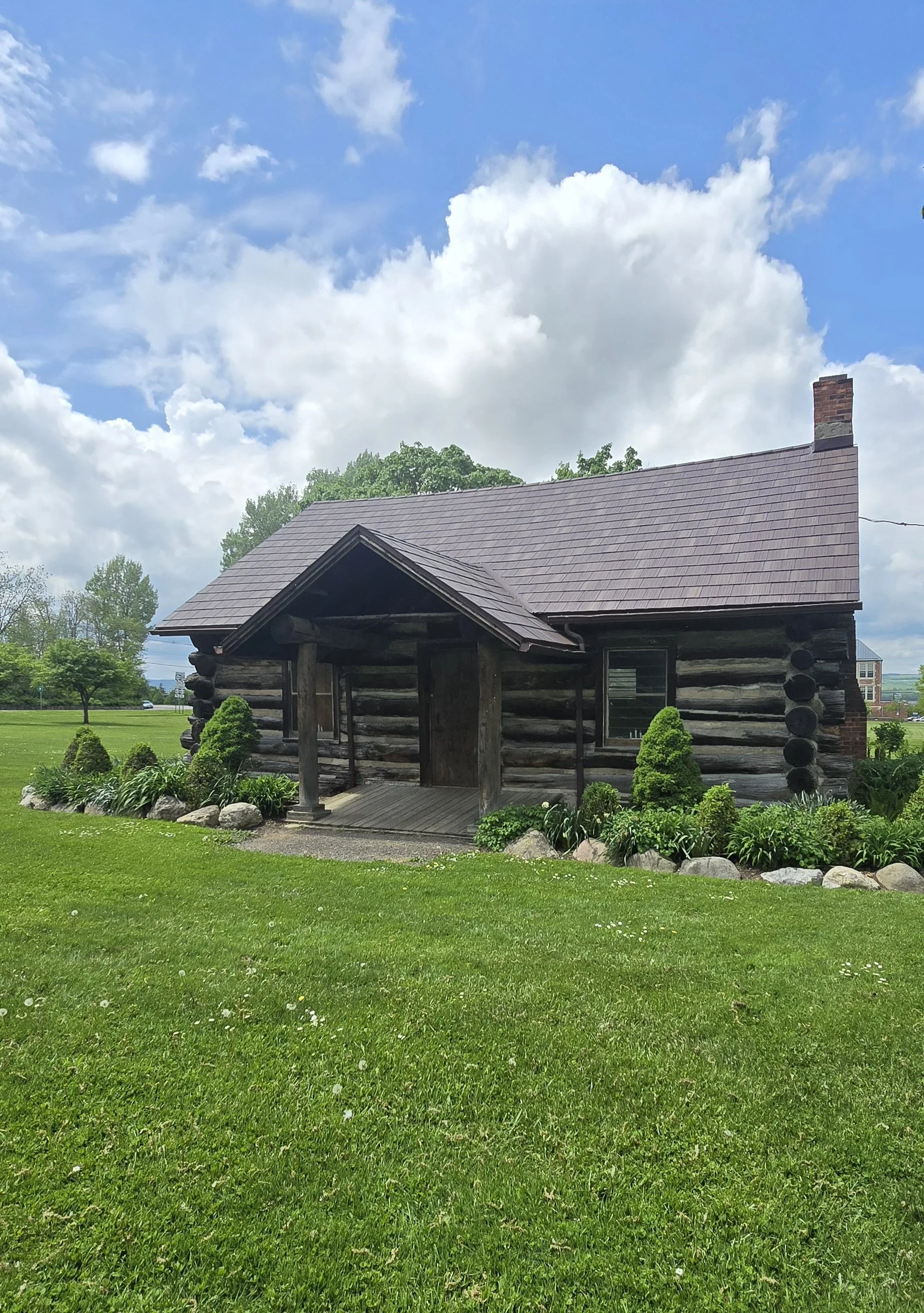 A rustic wooden log cabin with a shingled roof, set in a lush green yard under a partly cloudy sky, surrounded by landscaped plants and rocks.