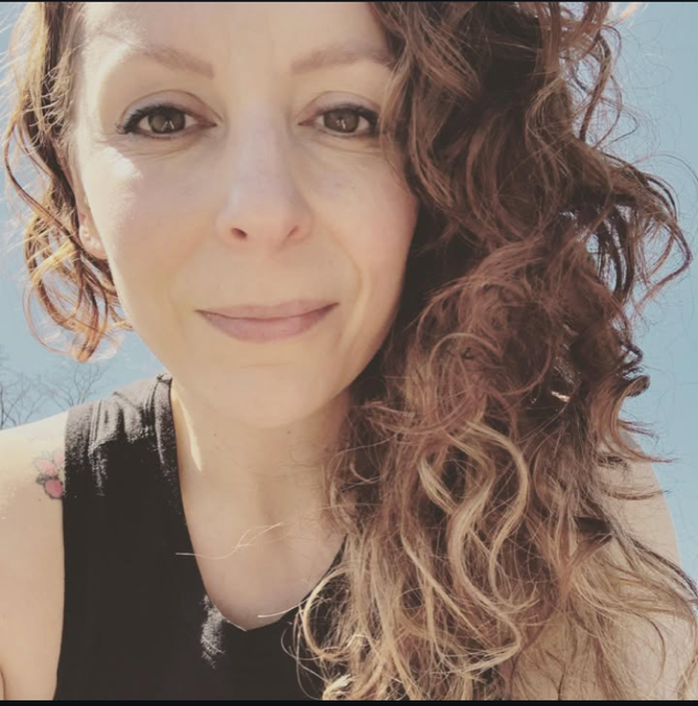 A woman with curly red hair takes a selfie outdoors in bright sunlight, with a clear blue sky in the background.