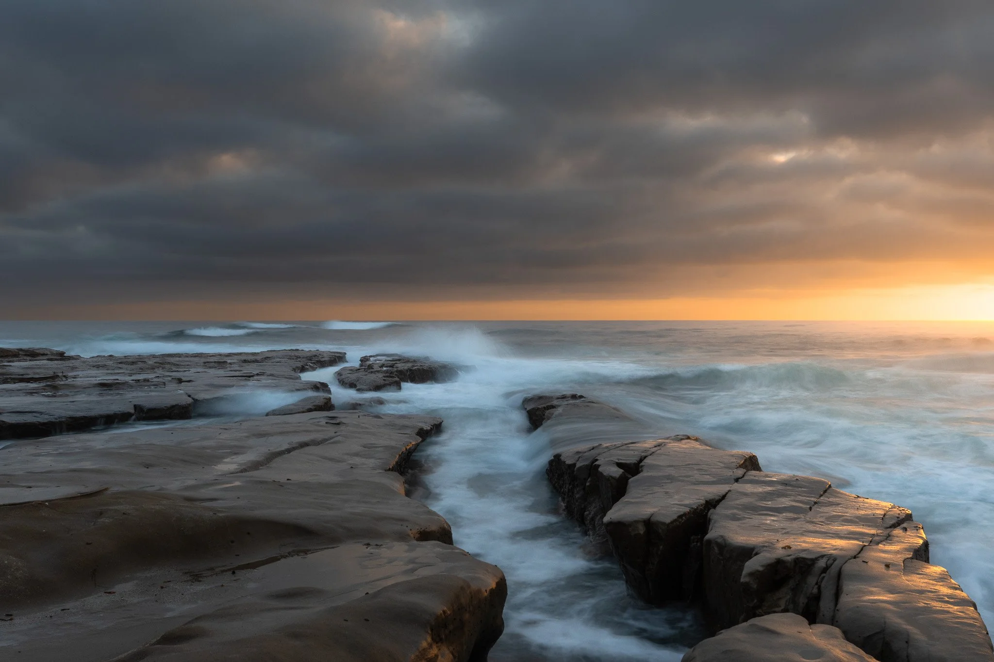 Sunset over a rocky beach with waves crashing against the rocks, dark clouds in the sky.