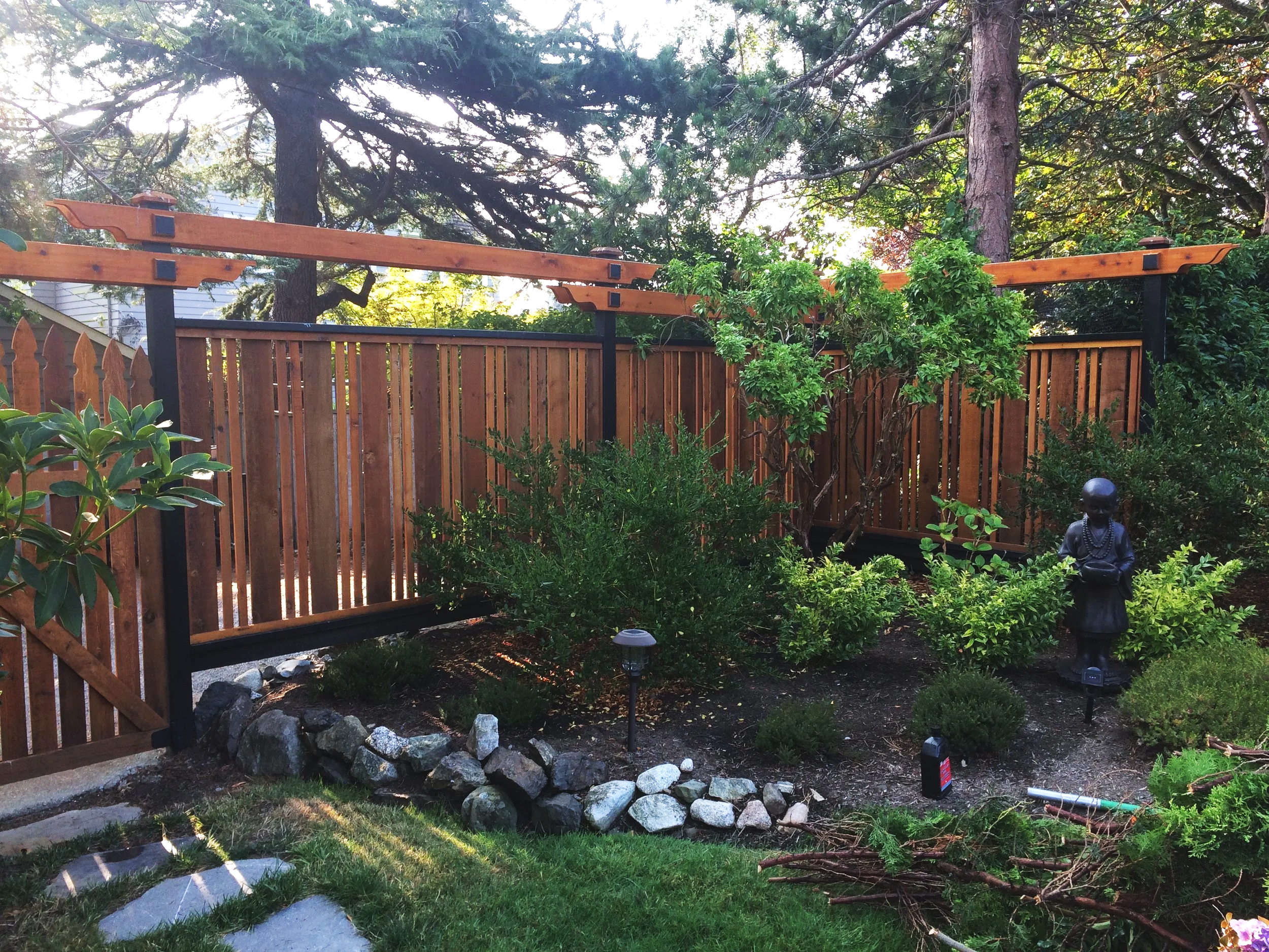 A backyard garden with a wooden fence, decorative plants, and a small statue of a child holding what appears to be a book or a tablet.