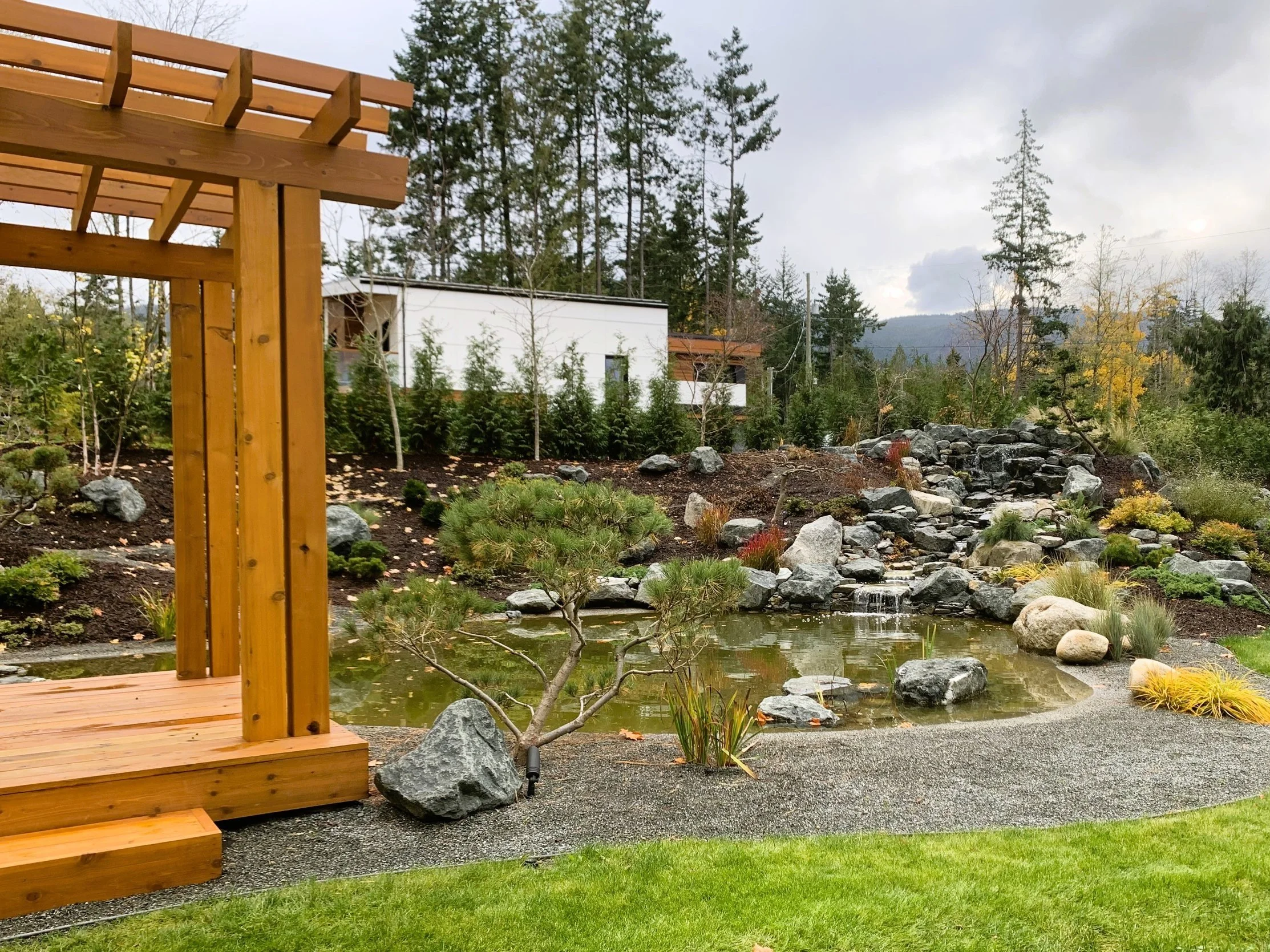 Backyard garden with a small pond, rocks, landscaped plants, a waterfall feature, and a wooden deck with steps, surrounded by trees and a modern white building in the background.