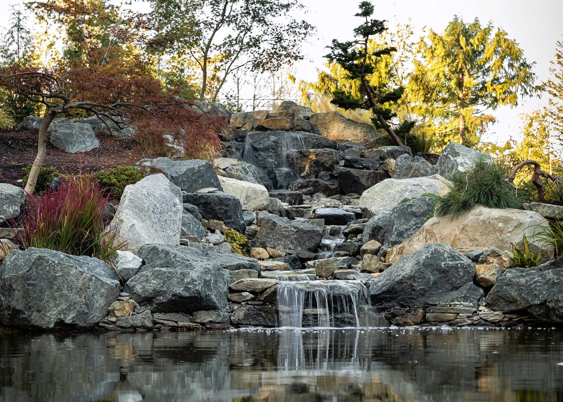 A small waterfall flows over rocks into a pond, surrounded by trees and plants.