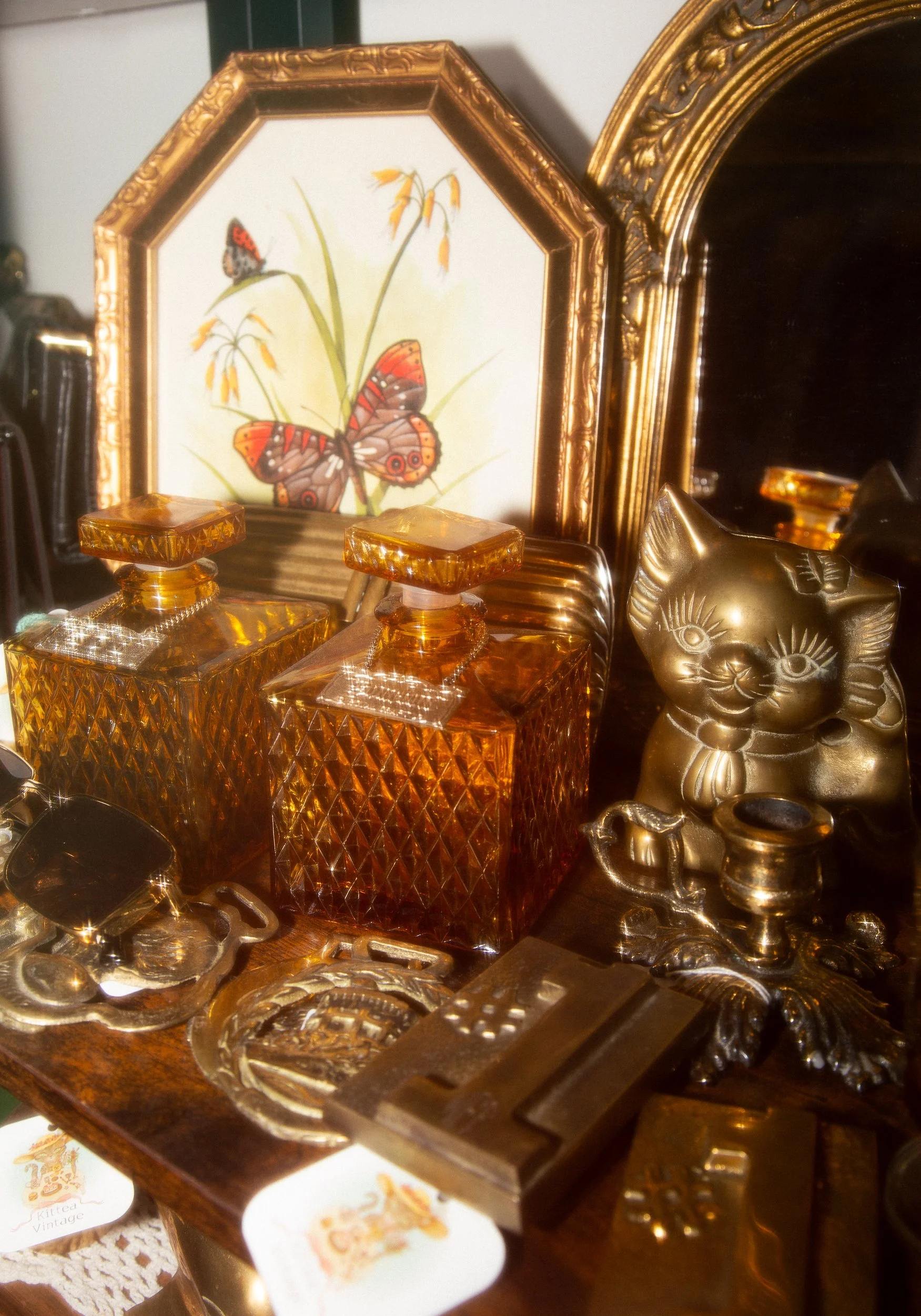 Decorative items on a wooden surface including amber-colored glass perfume bottles, a bronze Maneki-neko figurine, and various small decorative objects in front of a framed butterfly and plant illustration with a gold ornate mirror in the background.