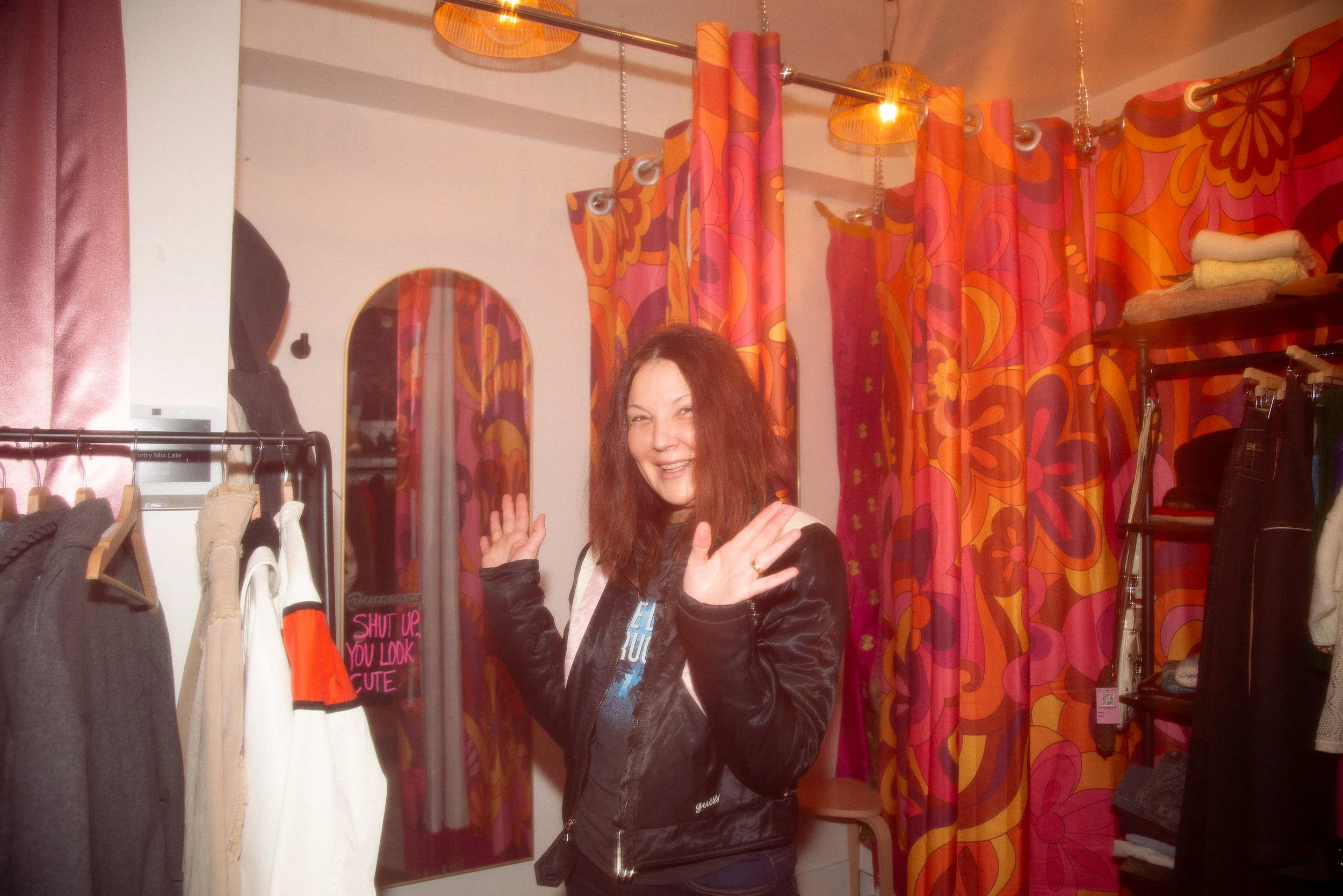 Woman smiling and posing with hands up in a boutique with floral curtains and clothing racks.