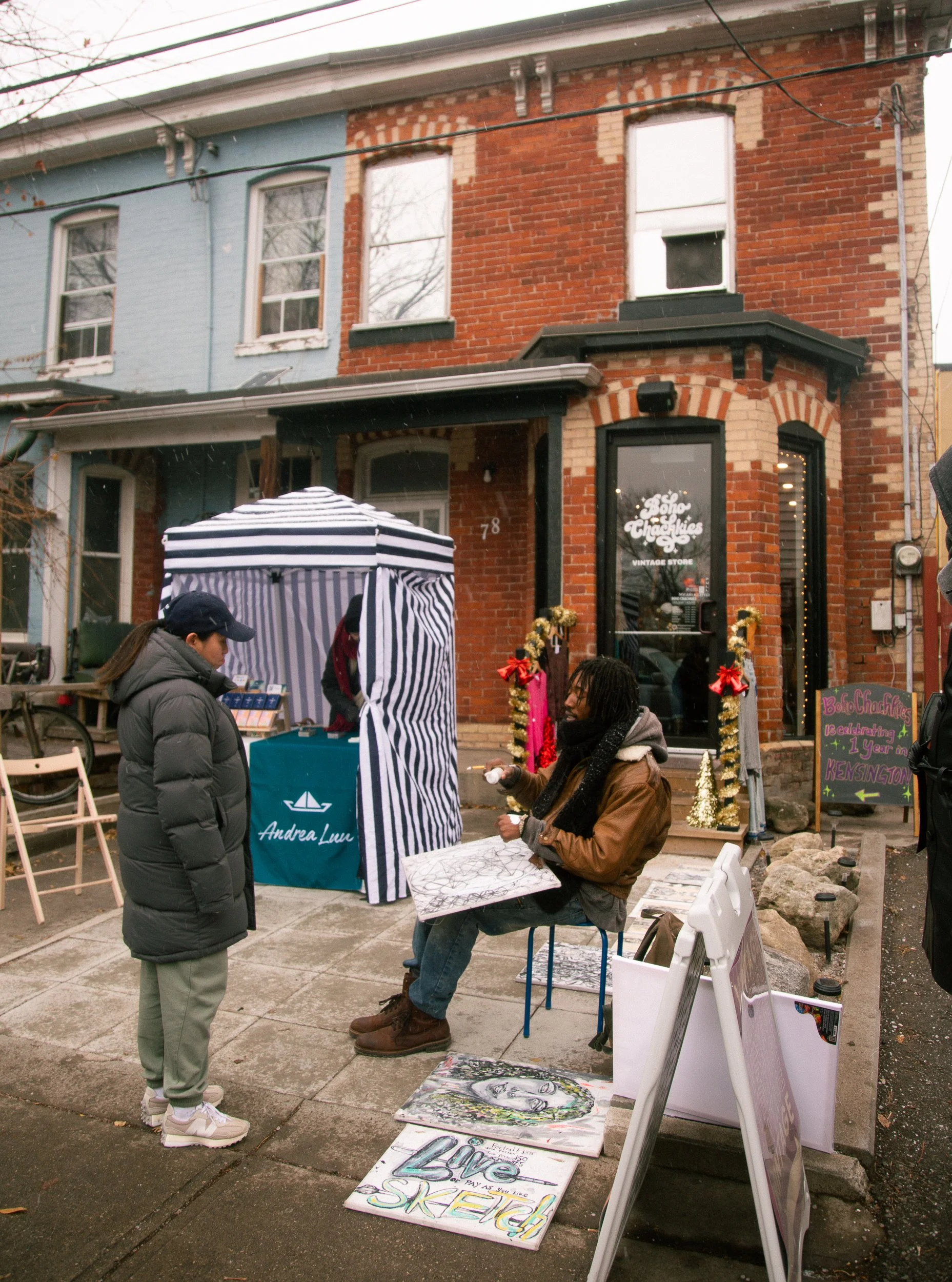 Man sitting on a chair displaying artwork for sale, talking to woman standing on sidewalk, street market scene in front of brick building with a vintage store sign, and decorated for the holidays.
