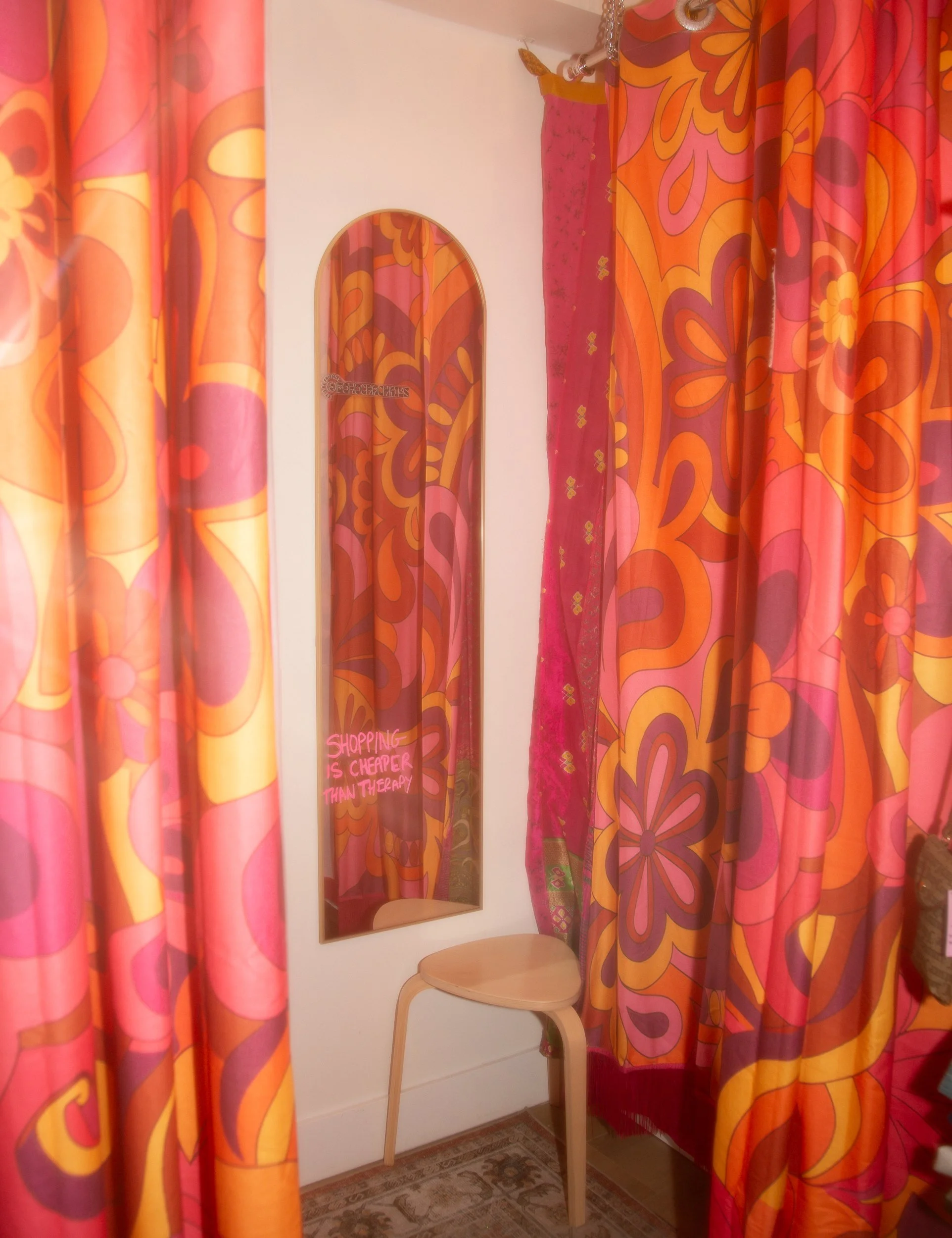 A fitting room with vibrant pink, orange, and purple floral-patterned curtains. A small wooden stool is positioned in front of a tall, arched mirror that has a pink sign on it reading 'Shopping is cheaper than therapy.' The room has a pink curtain wi