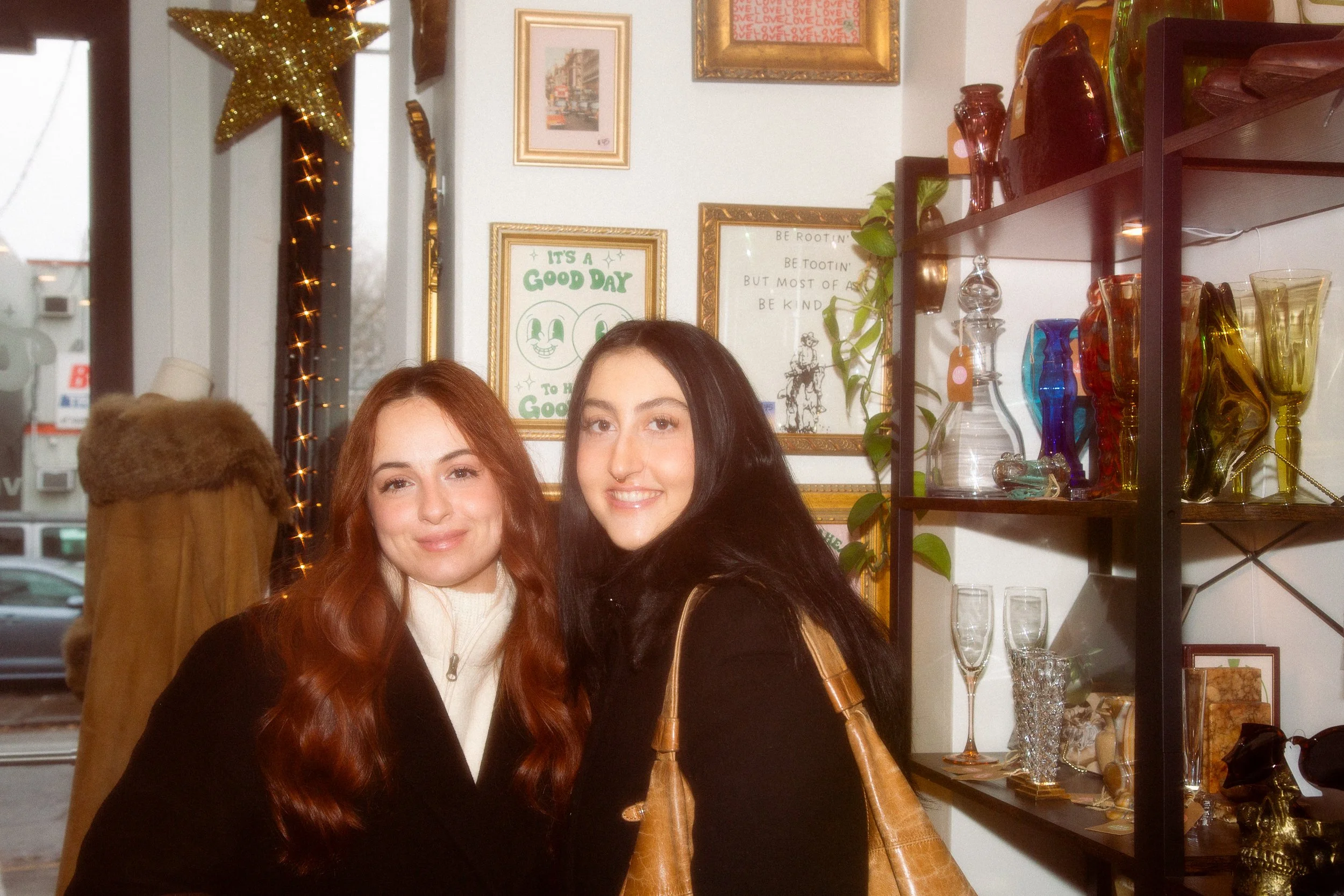 Two young women with long hair, one with red hair and the other with black hair, smiling and posing in an indoor setting that appears to be an art or gift shop, with framed pictures, colorful glassware, and decorative items on shelves in the backgrou
