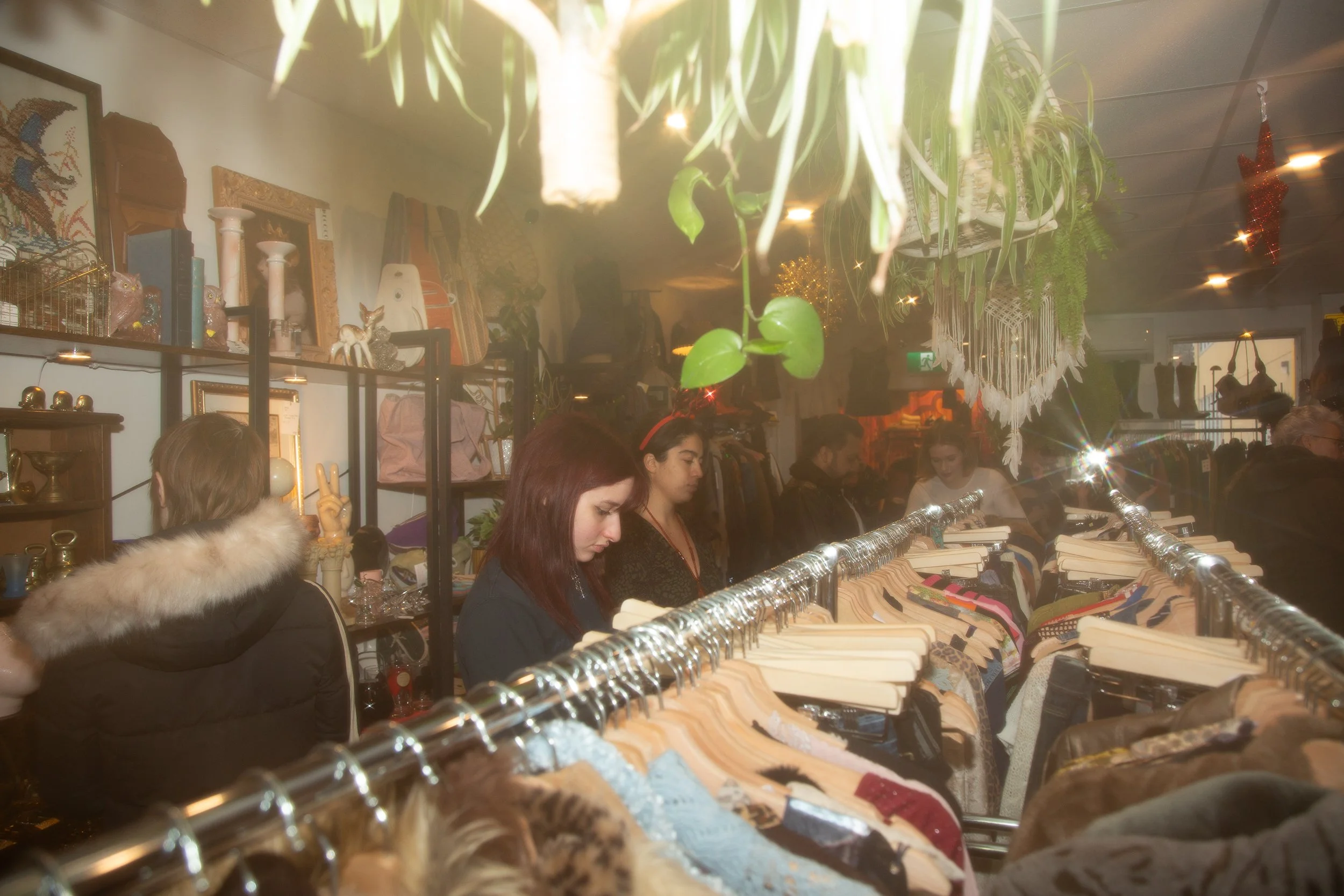 People shopping at a thrift store or flea market, browsing clothing on racks and looking at items on shelves, with clothing, decor, and plants visible in the background.