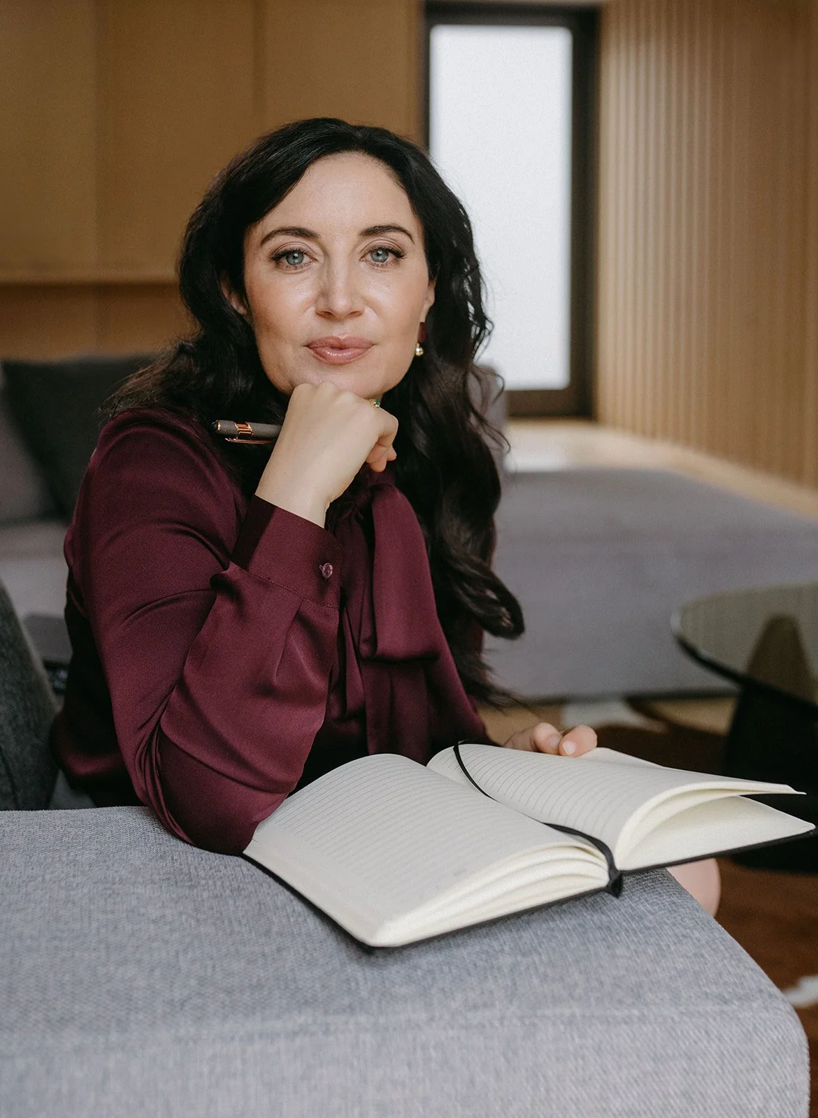 A woman with long dark hair and blue eyes sitting on a gray sofa with an open notebook in front of her, resting her chin on her hand with a pen, in a modern room with wood-paneled walls and a window in the background.