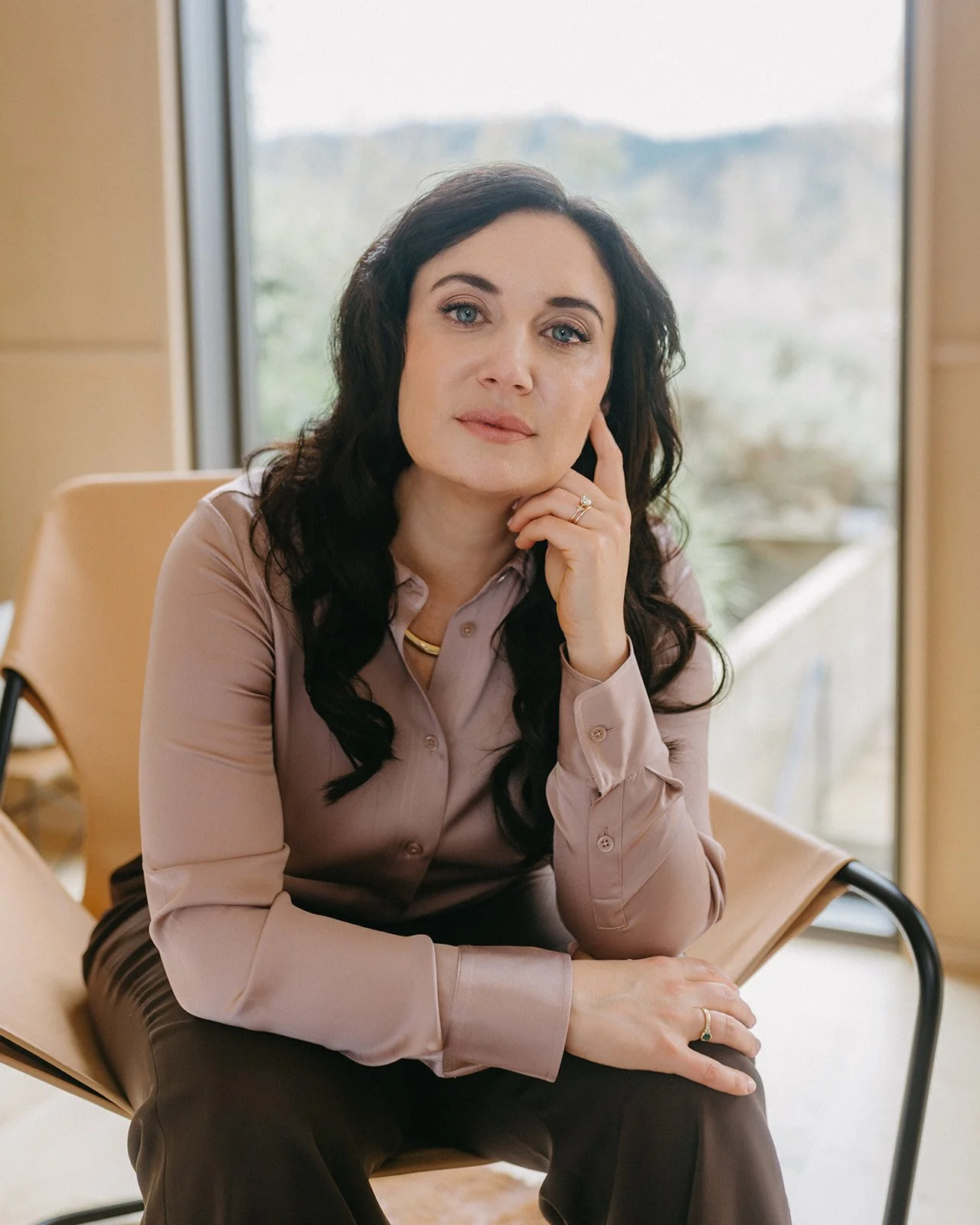 A woman with long dark hair, wearing a beige button-up shirt, seated indoors near a large window, looking at the camera with a slight smile.