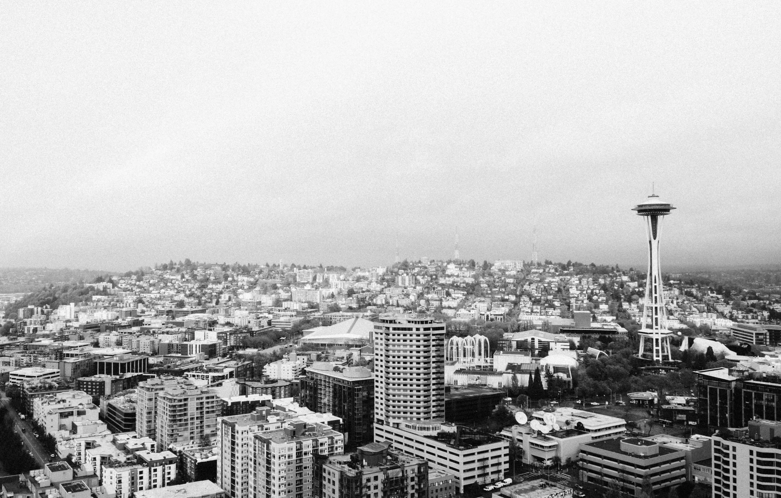 Black and white aerial photo of downtown Seattle, Washington, featuring the Space Needle skyscraper with city buildings, hills, and overcast sky in the background.