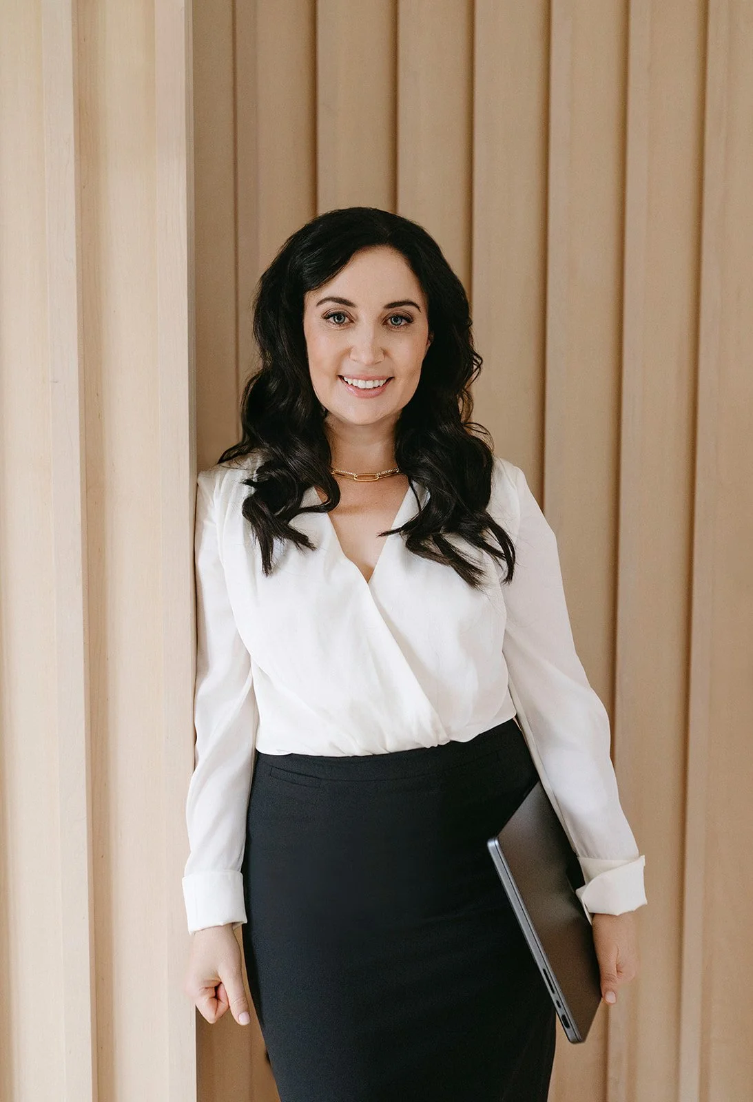 A woman with long dark hair wearing a white blouse and black skirt, holding a silver laptop, standing against a wooden wall background, smiling at the camera.