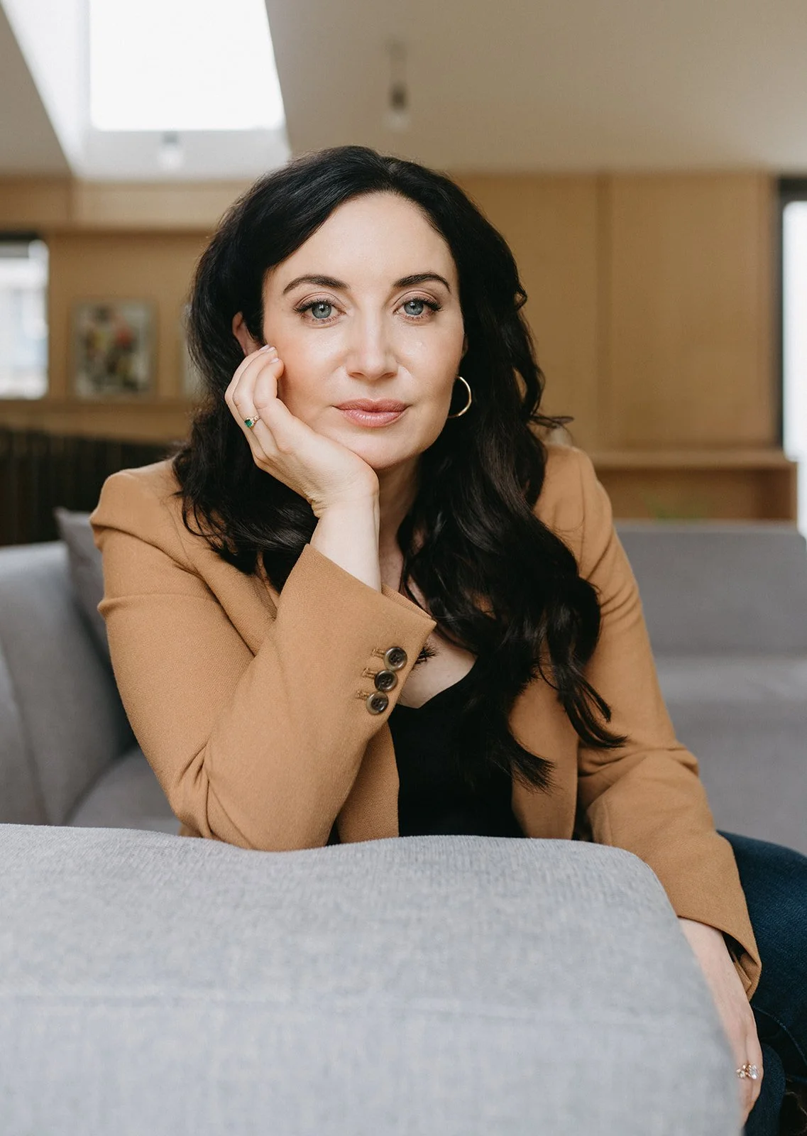 A woman with long dark hair and blue eyes, wearing a tan blazer, sitting on a sofa with her hand resting on her face, looking directly at the camera with a neutral expression in a well-lit indoor setting.