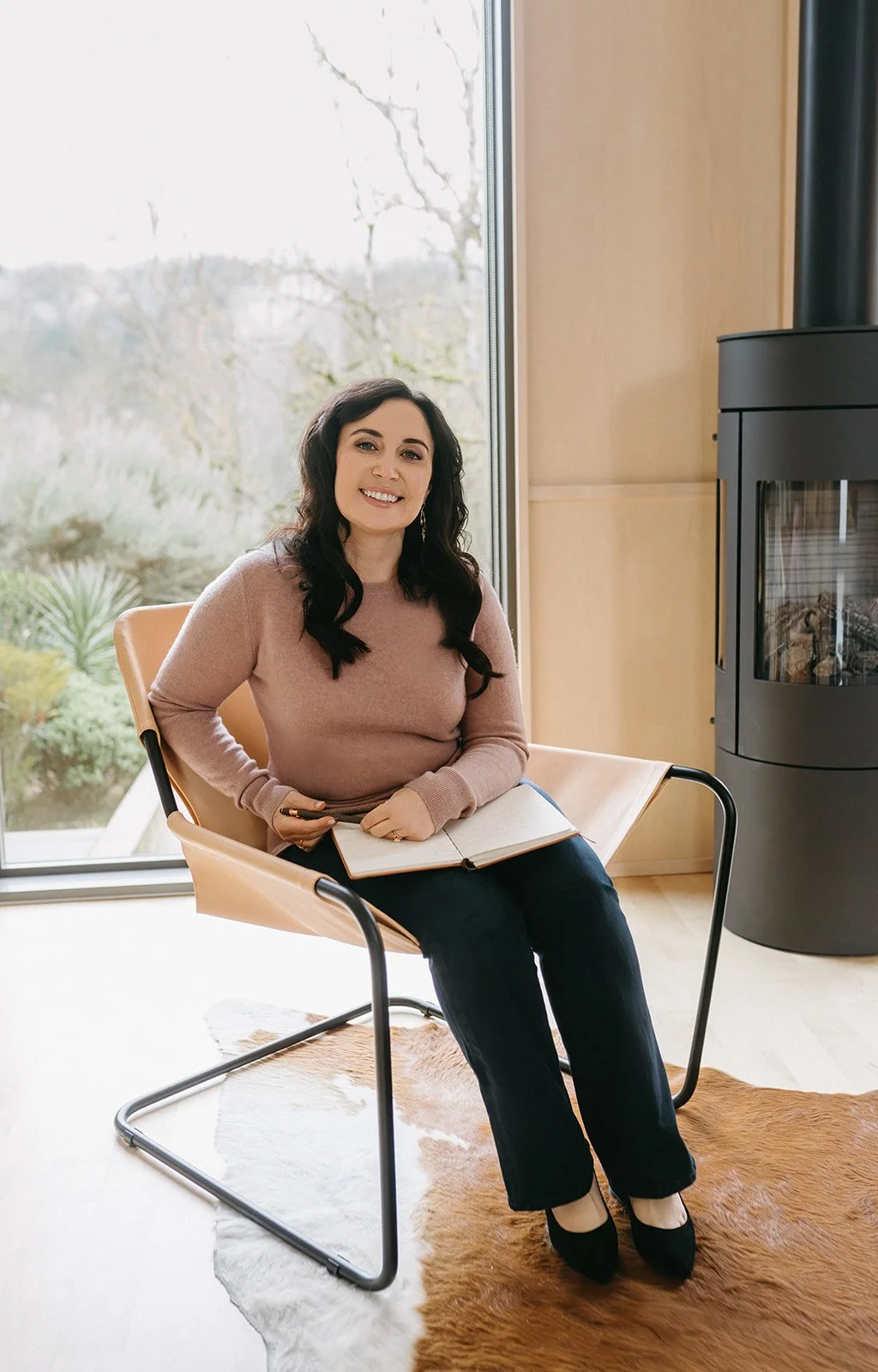 A woman with dark, curly hair smiling and sitting on a modern chair with a wooden frame, holding an open notebook and a pen, in front of a large window with greenery outside, next to a black wood stove, in a room with light wood walls and floors.