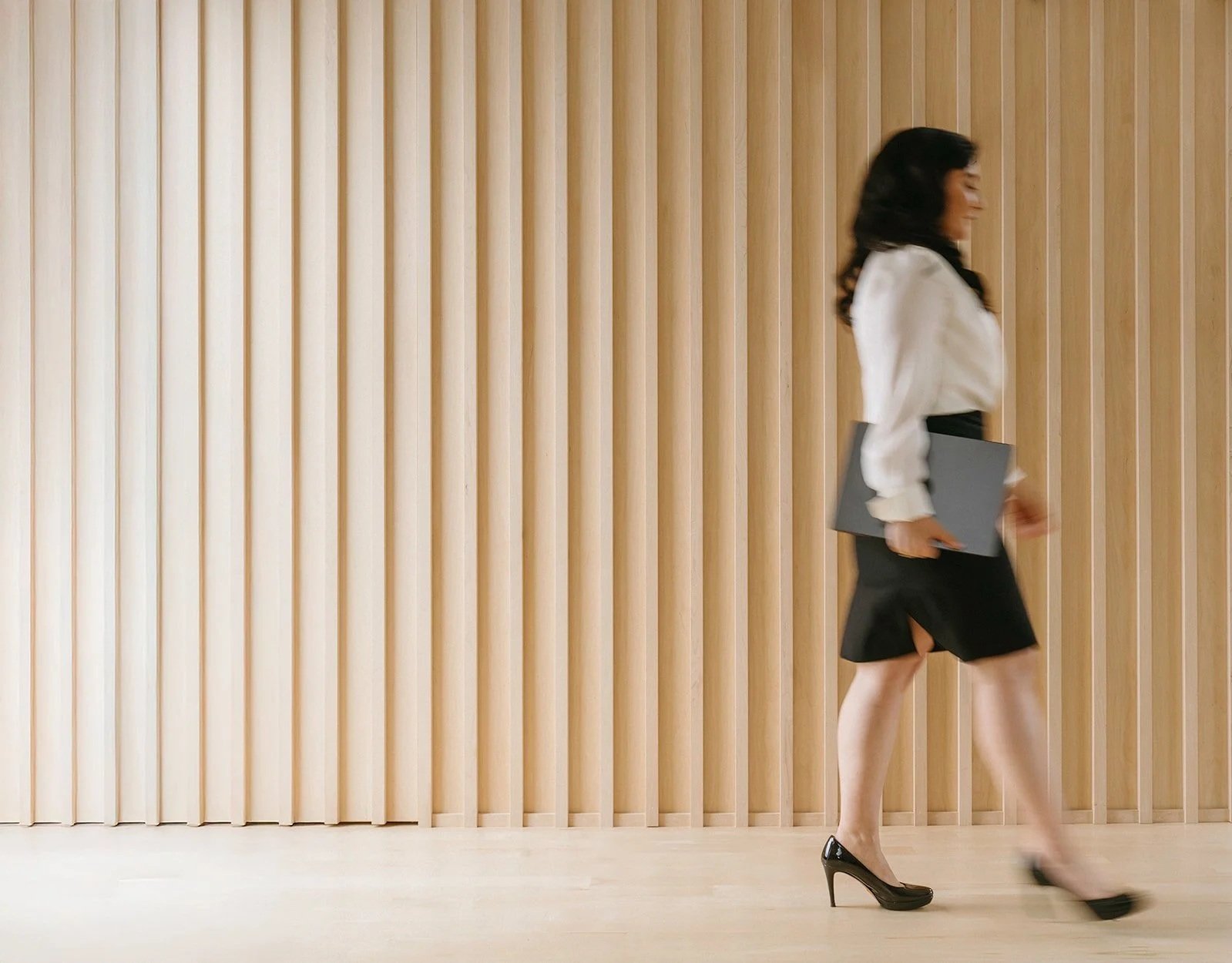A woman walking in front of a wooden slat wall holding a gray folder, wearing a white blouse, black skirt, and high heels.