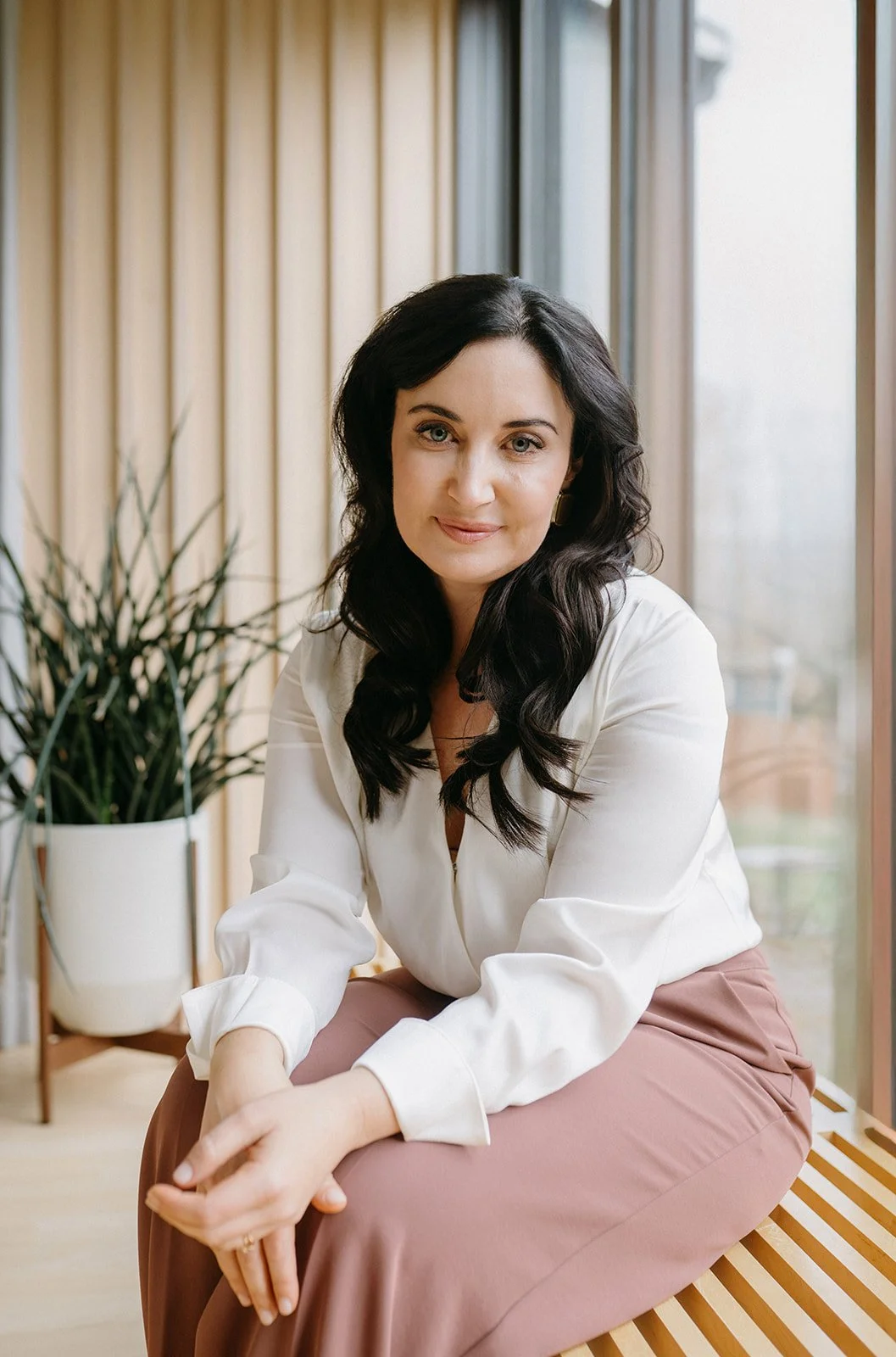 A woman with dark curly hair, wearing a white blouse and pinkish-brown pants, is sitting on a wooden bench, posing indoors near large windows with beige curtains and a potted plant nearby.