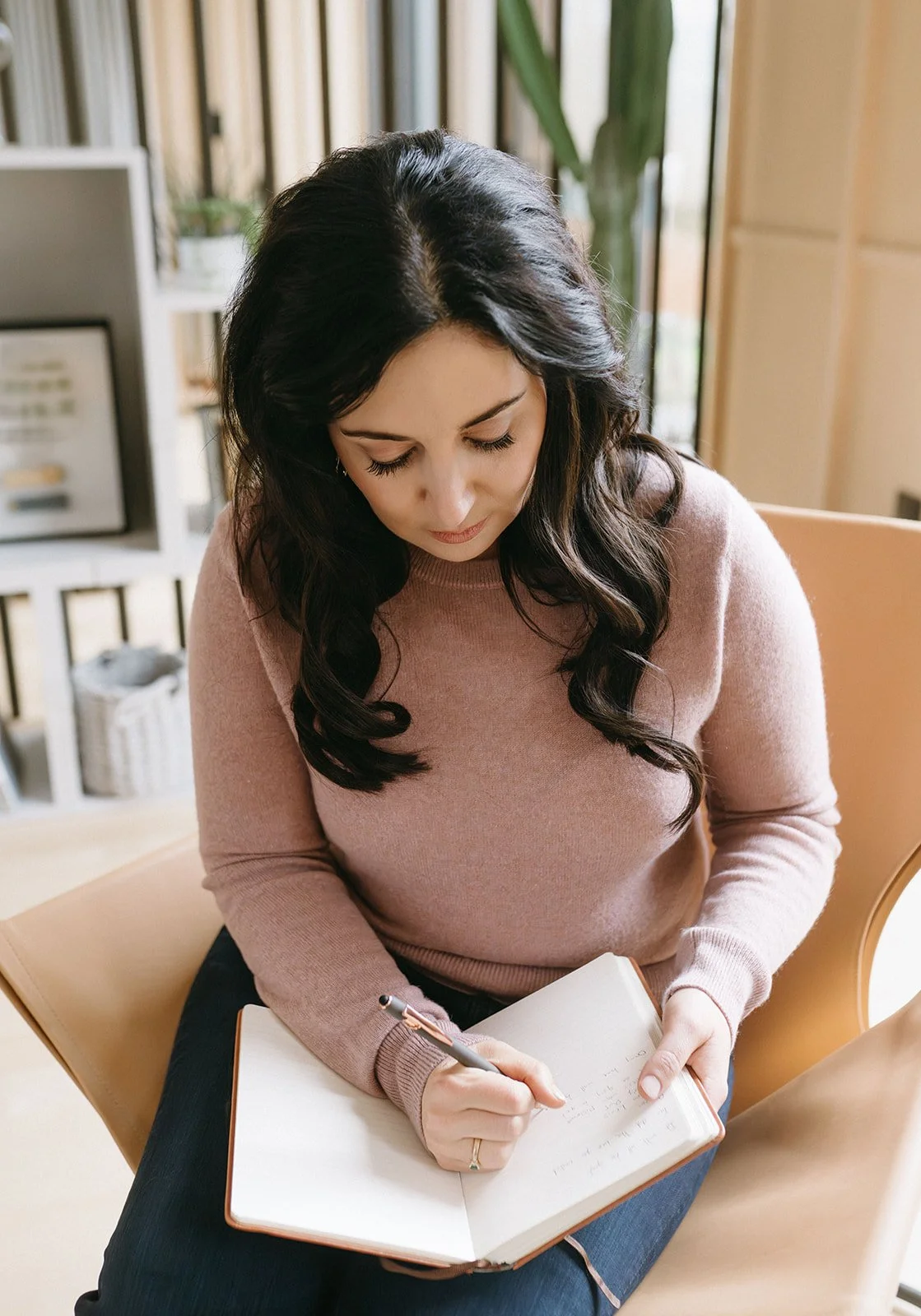 A woman with dark wavy hair seated and writing in a notebook with a pen, wearing a pink sweater, in a cozy, well-lit room.