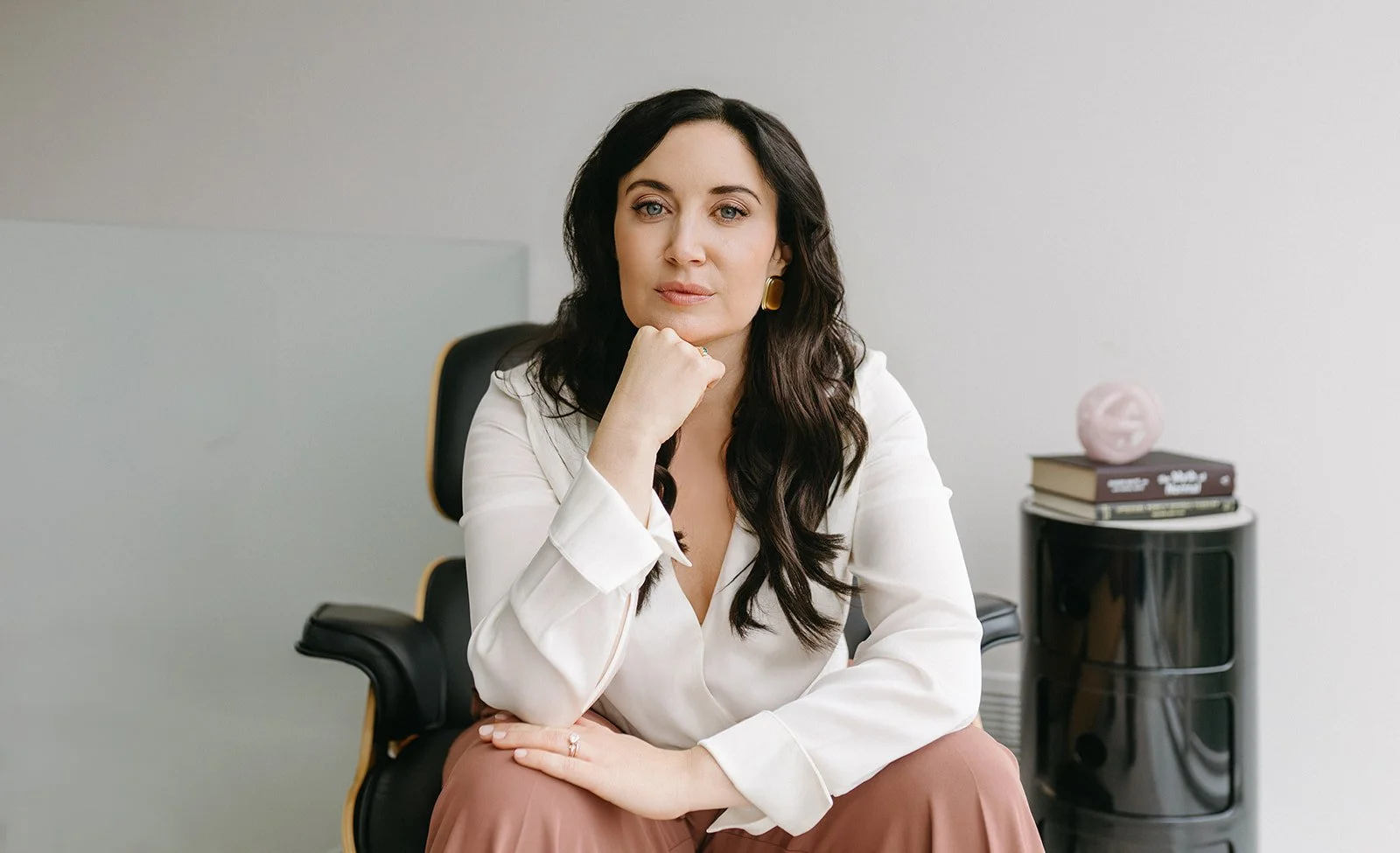 A woman with long dark hair sitting in an office chair, resting her chin on her hand, with a neutral expression, wearing a white blouse and brown pants, in a modern office setting.