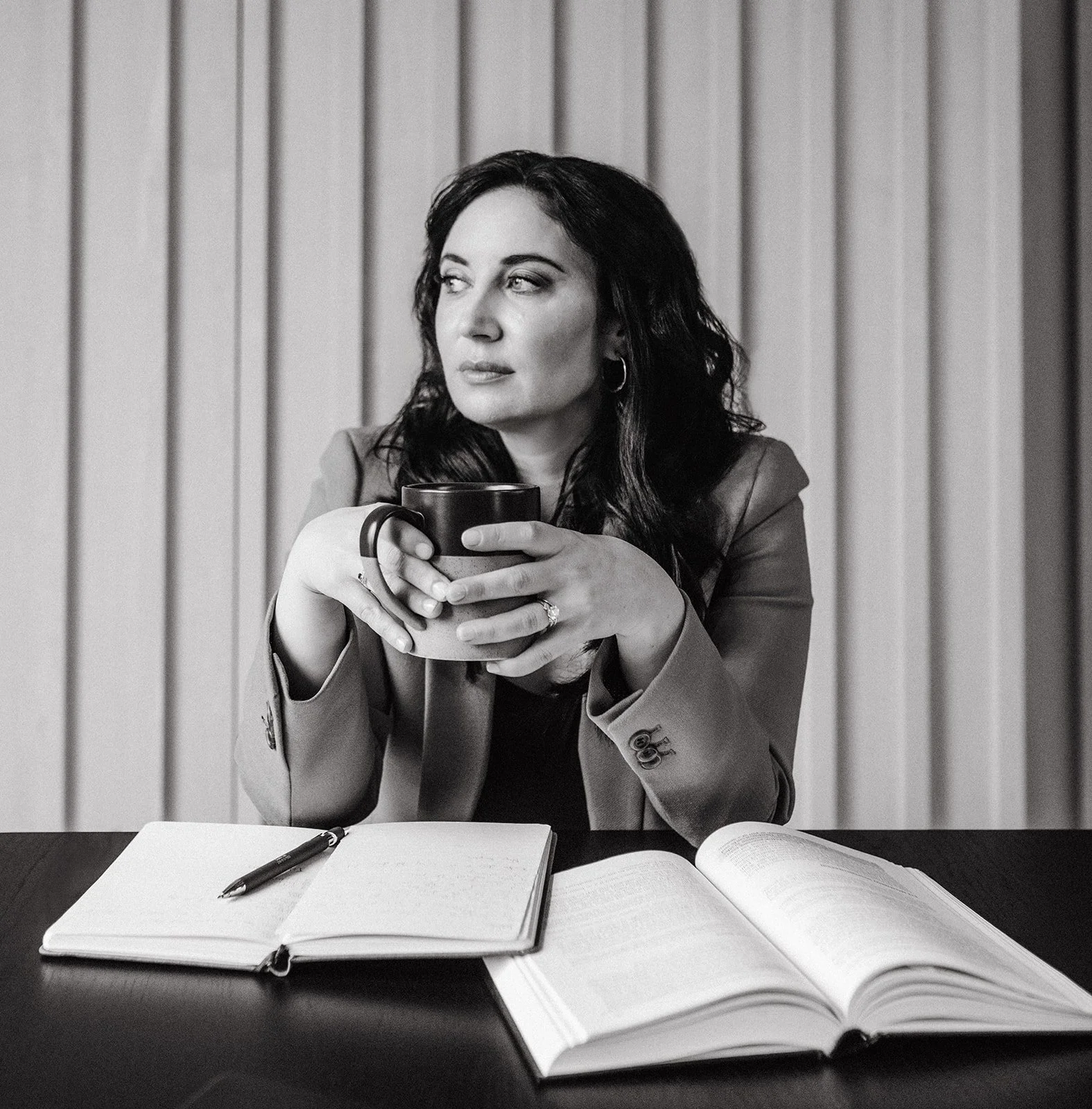 A woman sitting at a table holds a coffee mug, with an open notebook, pen, and additional books on the table in front of her, in a setting with vertical gray paneling.