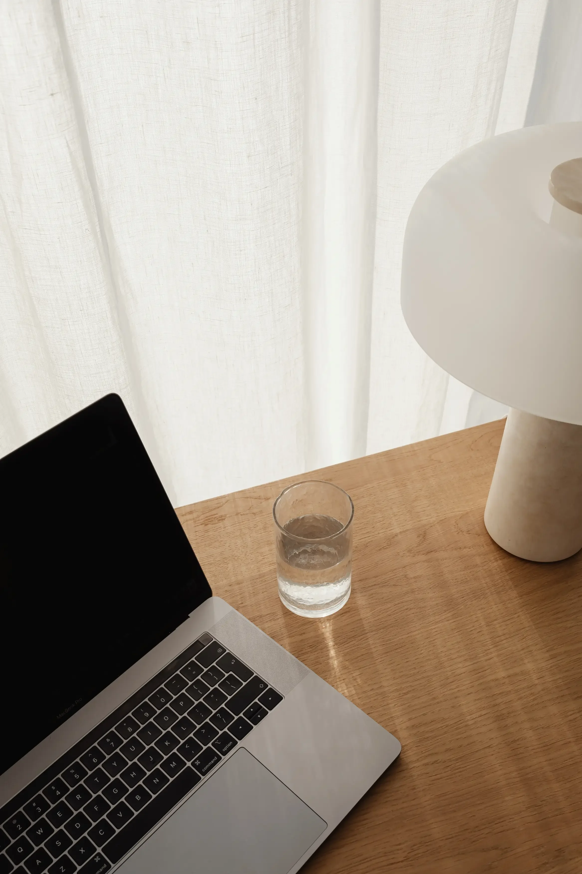 A laptop on a wooden desk with a glass of water, a white decorative vase, and white curtains in the background.