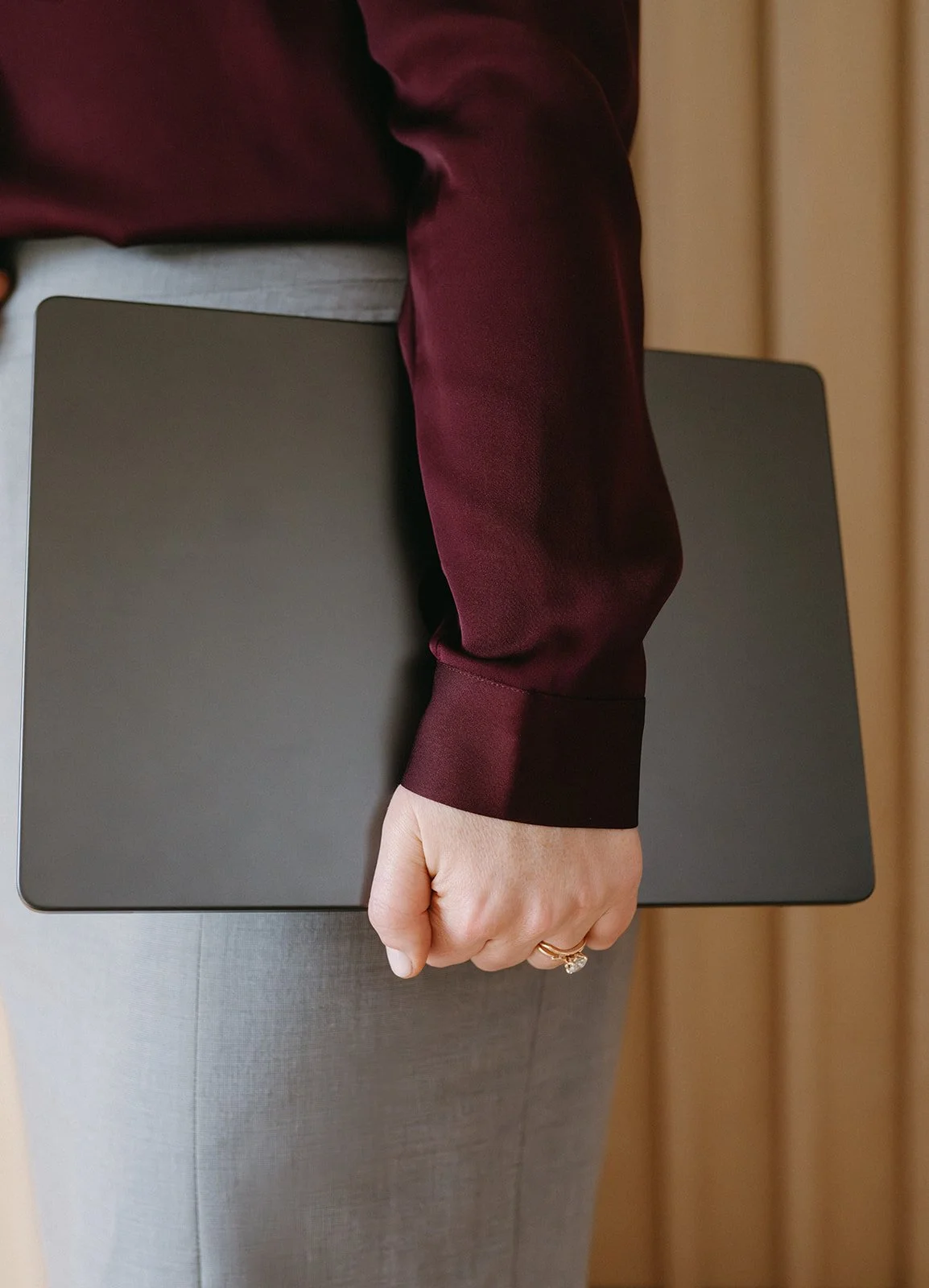 A person holding a closed laptop, wearing a maroon satin blouse, gray pants, and a ring on their finger.