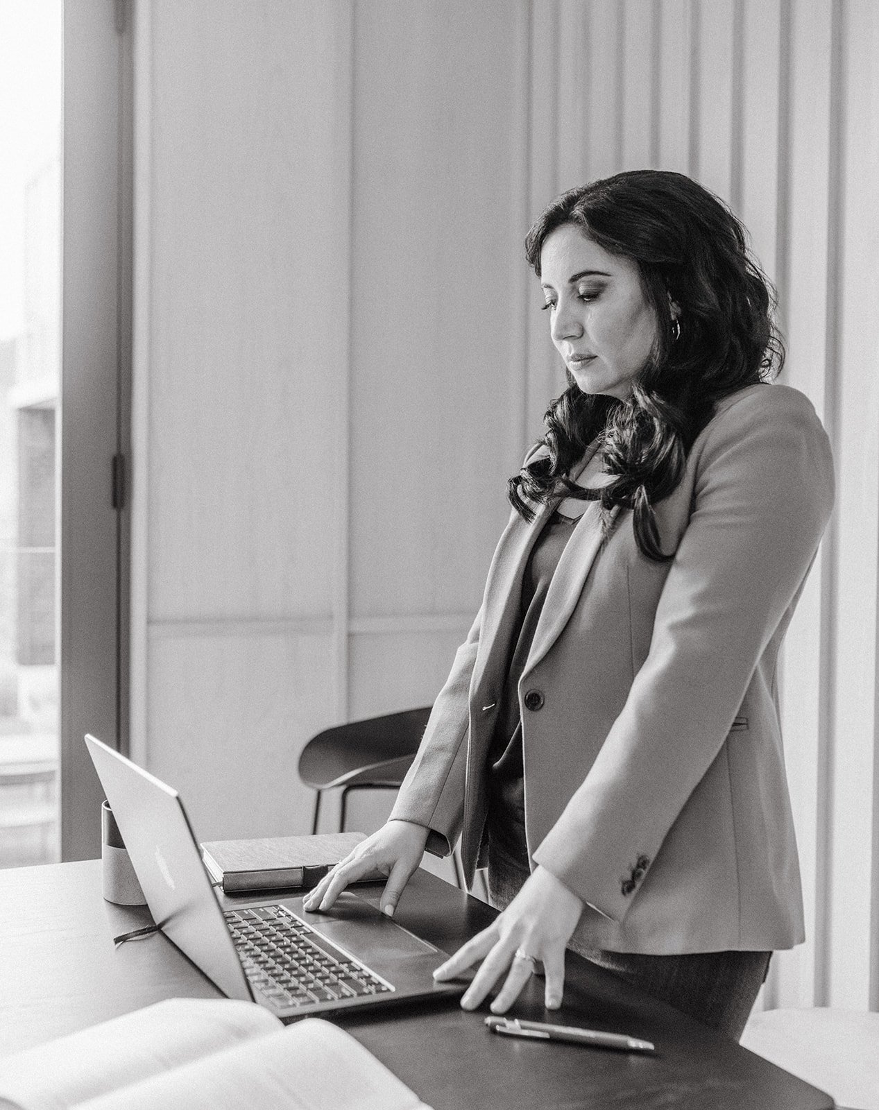 Black and white photo of a woman standing next to a laptop at a conference table, with papers and a pen, in an office setting with wood panel walls and a window.
