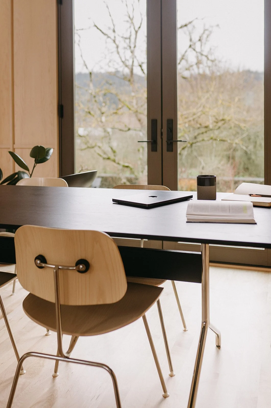 Interior of a modern dining or meeting area with a wooden table, chairs, laptop, an open notebook, a closed notebook, a black and white cup, and a potted plant, against large glass doors showing a view of trees outside.