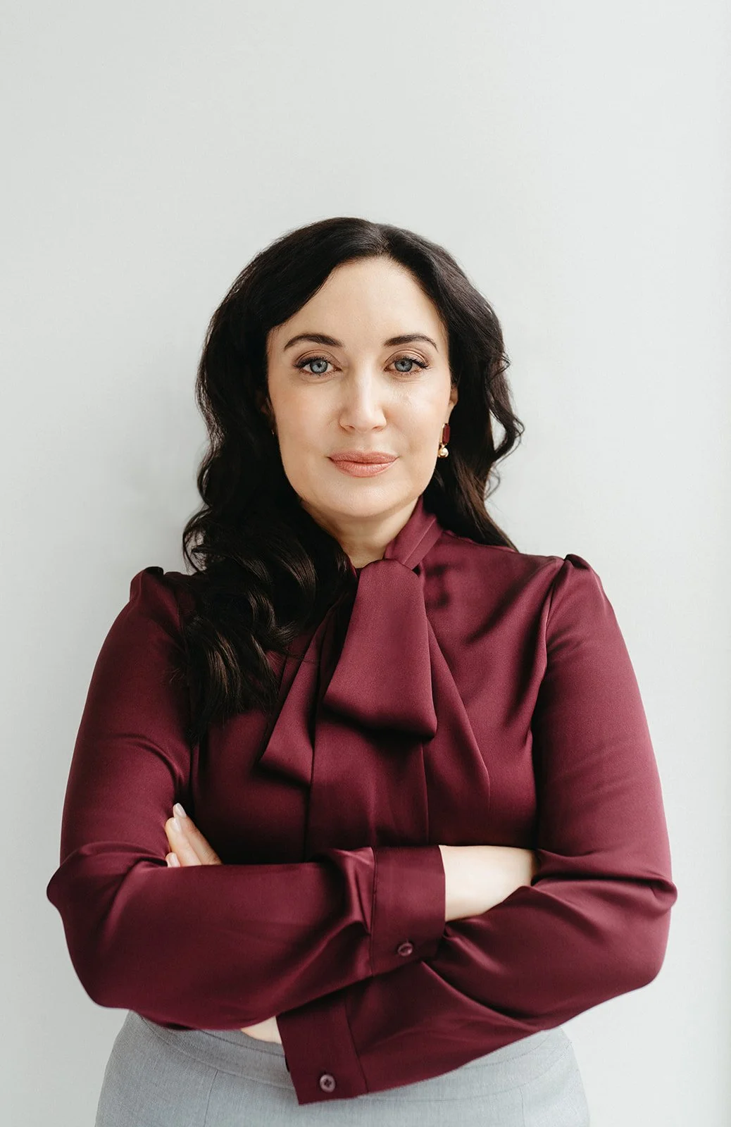 A woman with dark wavy hair and blue eyes, wearing a maroon blouse with a bow tie and gray pants, standing against a plain white background.