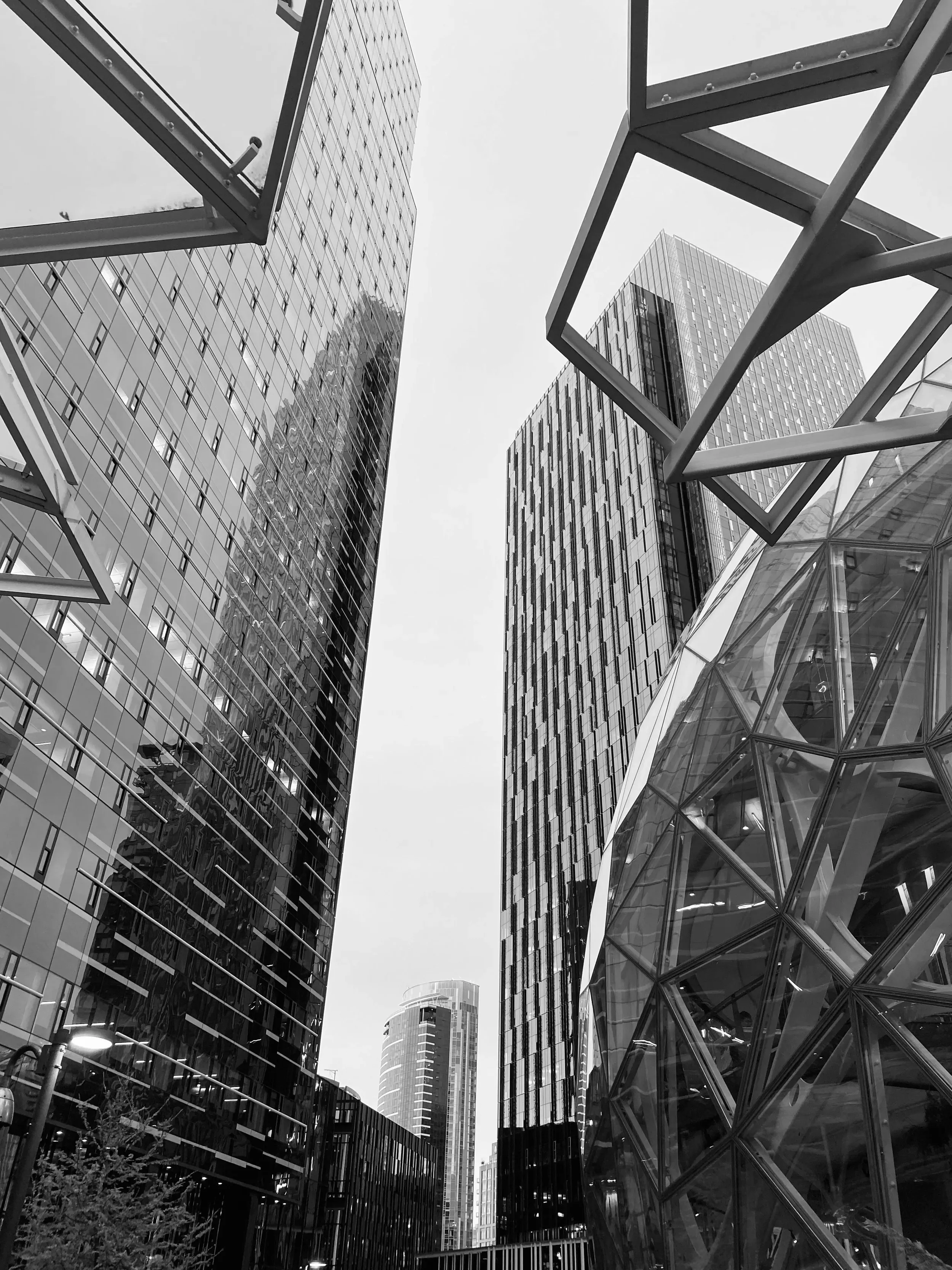Black and white photo of modern skyscrapers with glass facades, shot from street level looking upward, including architectural features like a glass pyramid structure on the right.