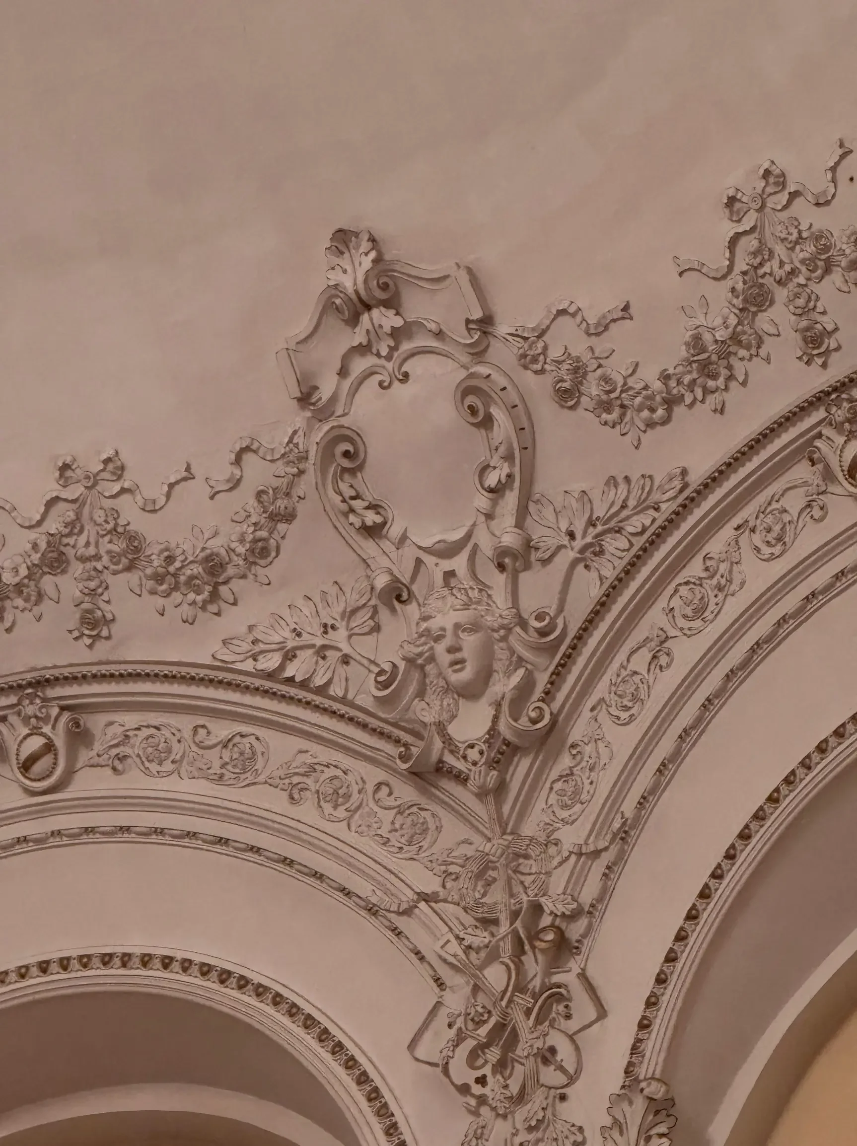 Intricate ceiling design with floral motifs, scrollwork, and a sculpted face of a woman, all painted in a classical style.