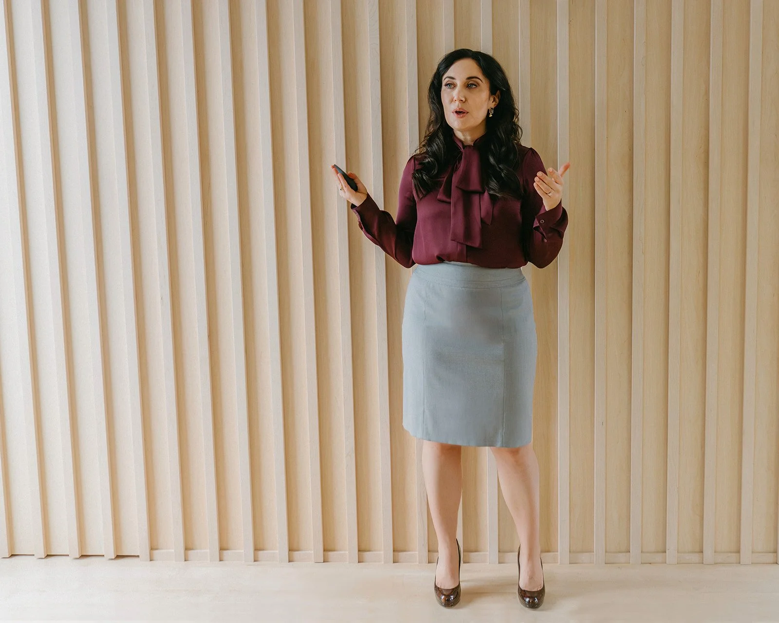 A woman in business attire stands in front of a wooden slat wall, wearing a burgundy blouse and a gray pencil skirt, holding a smartphone and speaking.