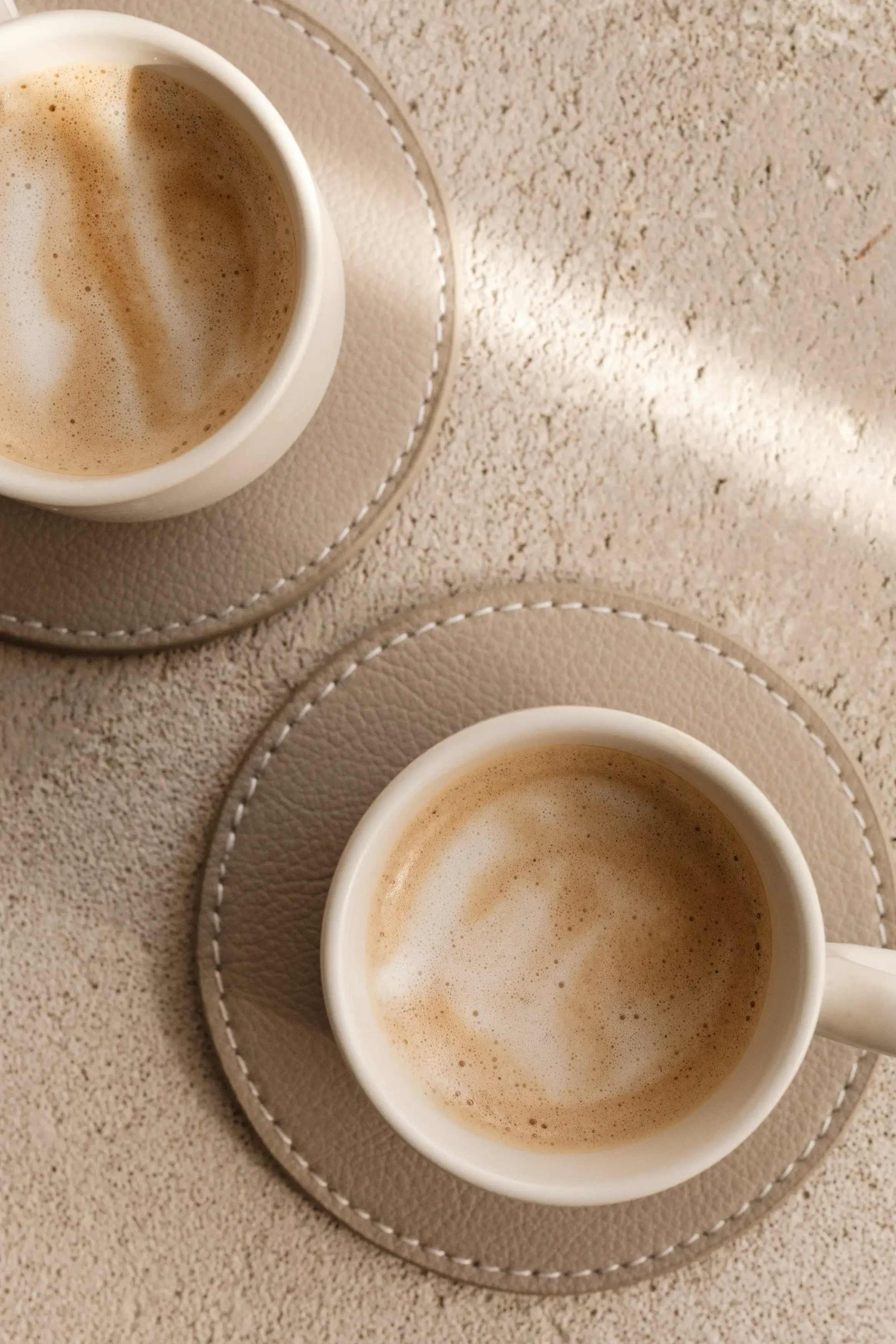 Two white cups of coffee with foam on top, placed on round beige leather coasters with white stitching, on a textured beige surface.
