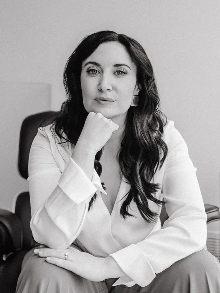 A black and white photo of a woman with long dark hair sitting in an office chair, resting her chin on her hand and looking at the camera.