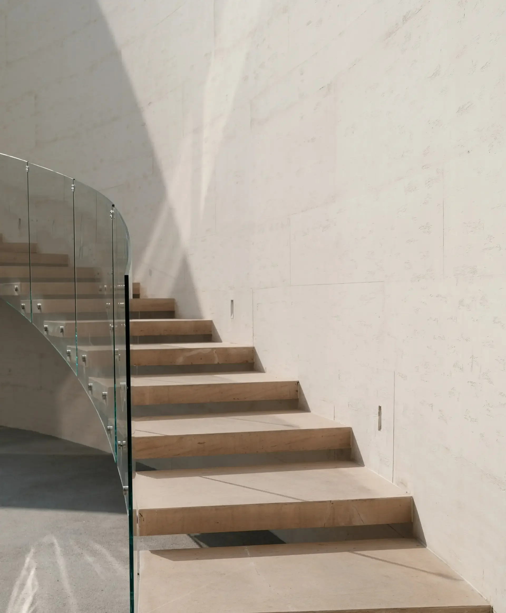 Interior view of a staircase with wooden steps and a glass railing, against a plain light-colored wall.