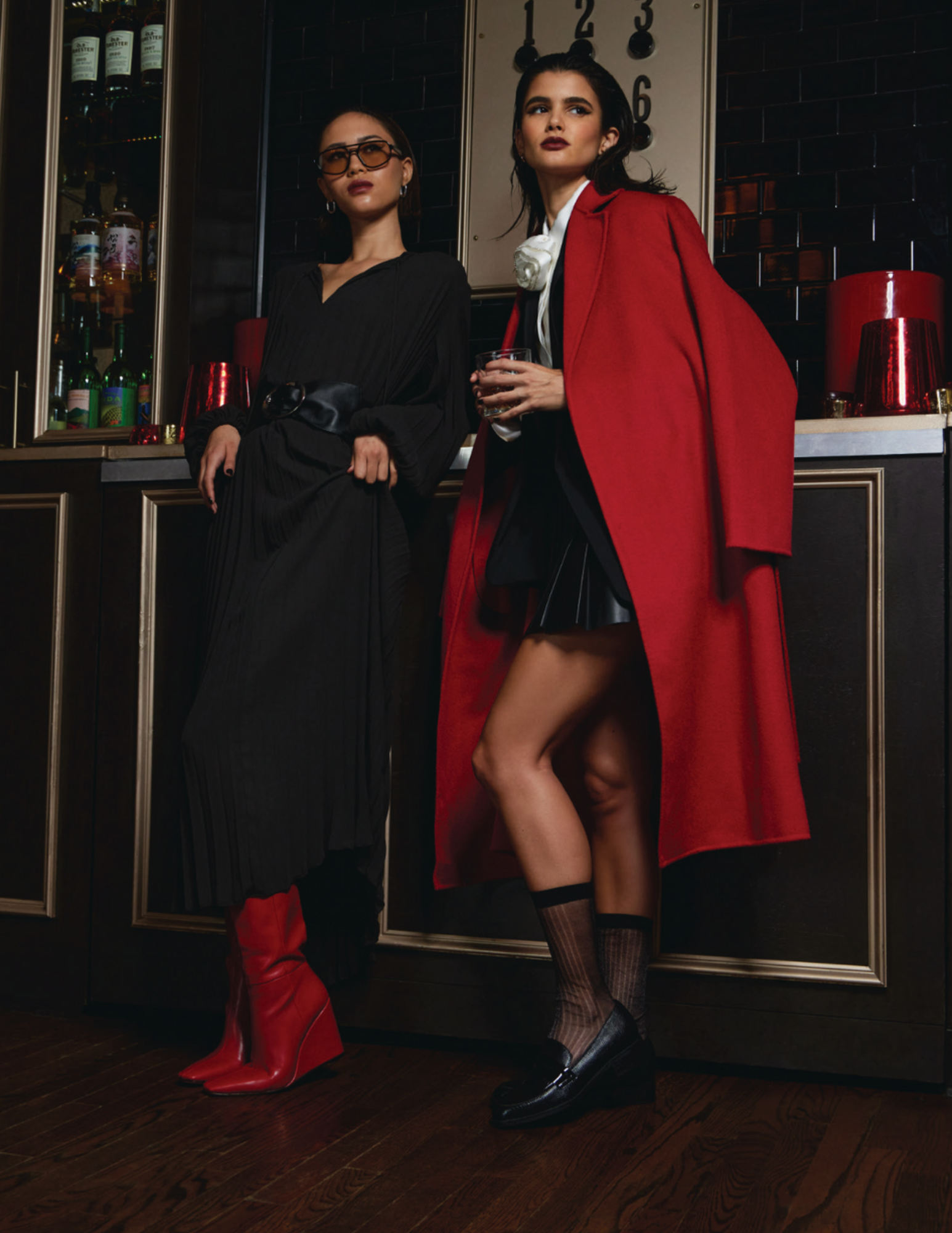 Two women in fashionable outfits standing in a bar or lounge area with liquor bottles and red decor in the background.