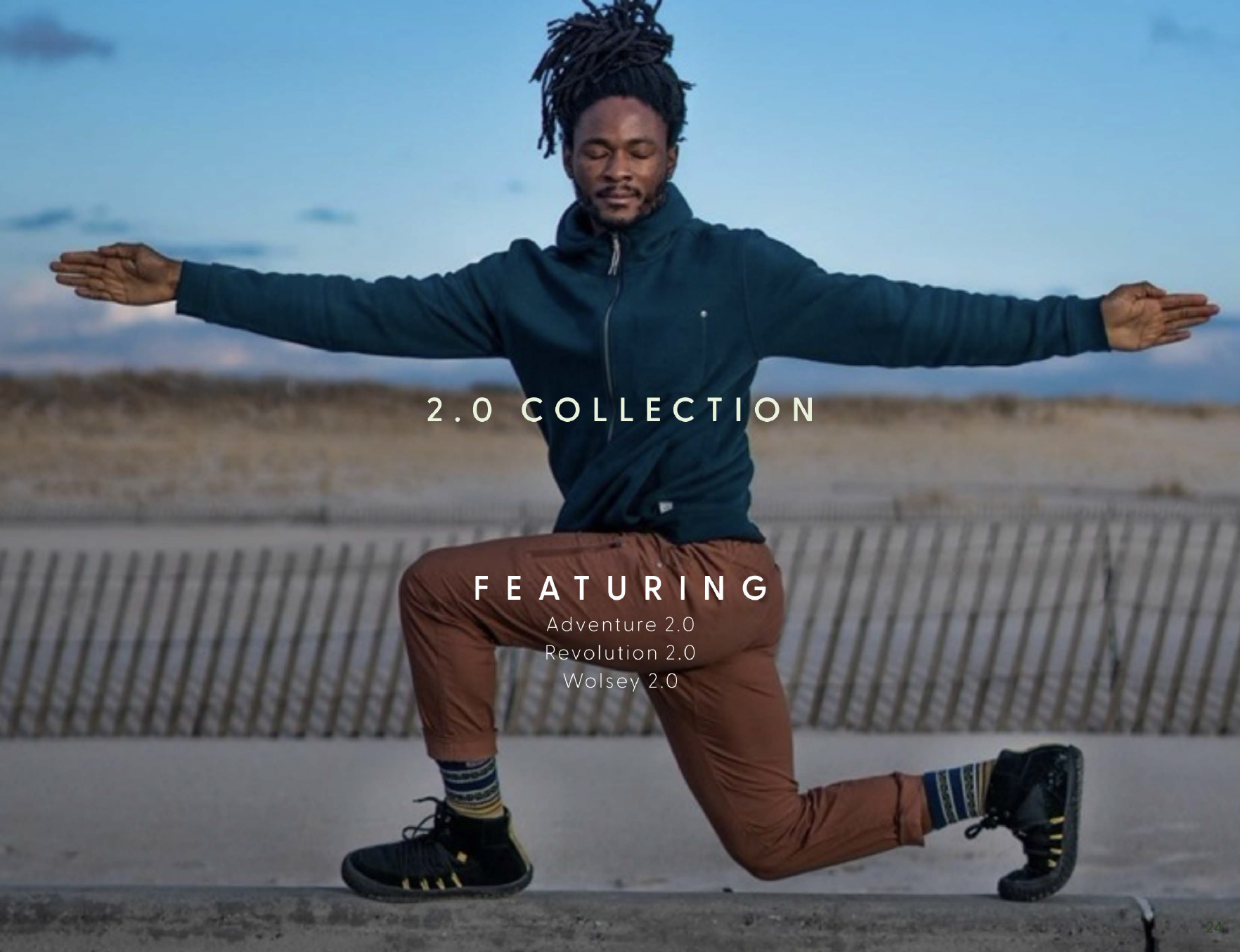 A man with dreadlocks is doing a yoga pose outdoors on a concrete path, wearing a blue jacket, orange pants, and striped socks with black shoes. The background shows a barrier and a landscape with a cloudy sky.