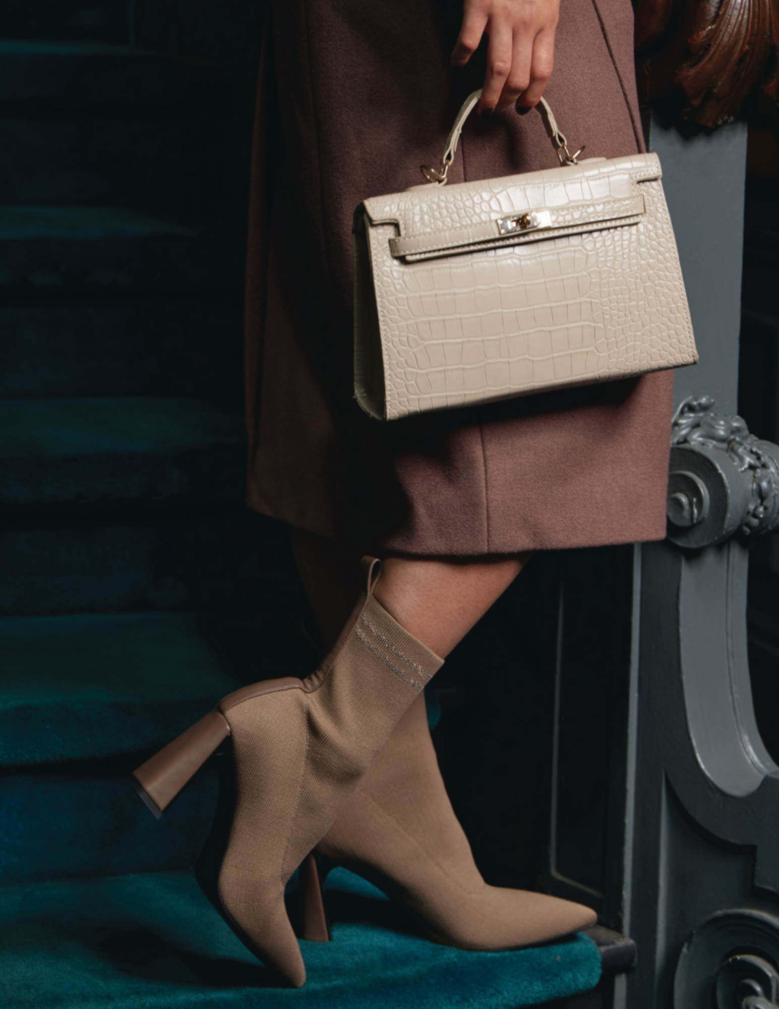Person holding a beige crocodile-embossed handbag standing on a staircase, wearing beige high-heeled boots and a brown coat.