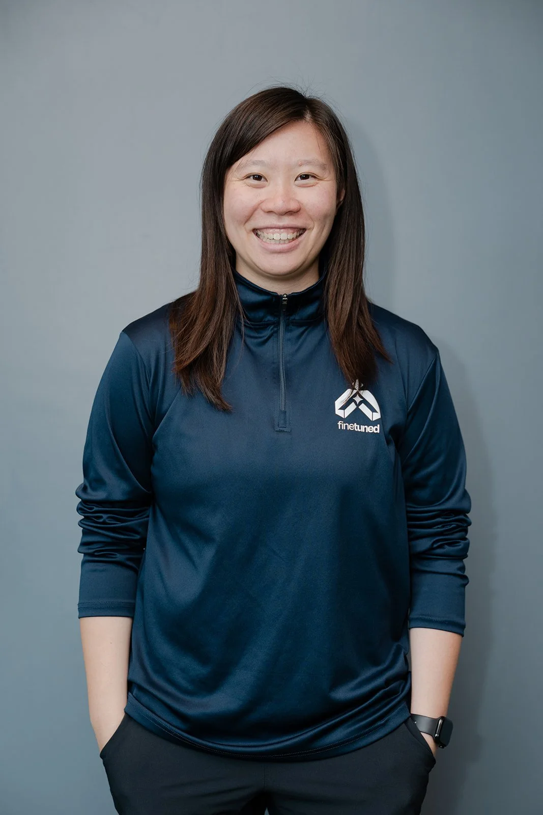 A smiling woman with long dark hair wearing a navy blue sports jacket with a logo that says 'finetuned' on it, standing against a plain gray background.