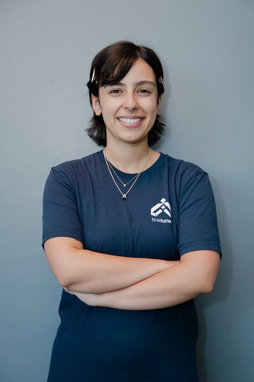 Close-up of a young woman with long brown hair, wearing a navy blue t-shirt with the 'finetuned' logo, silver necklaces, and a nose ring, smiling against a light blue wall.
