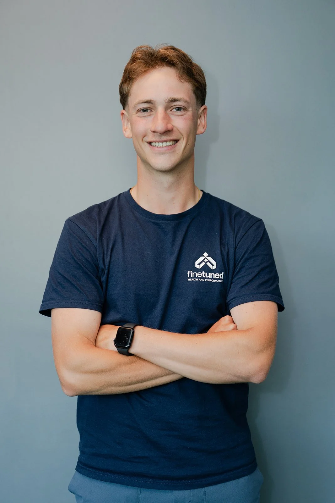 Young man smiling, wearing a black athletic T-shirt with 'finetuned' logo and text, standing against a plain gray wall.