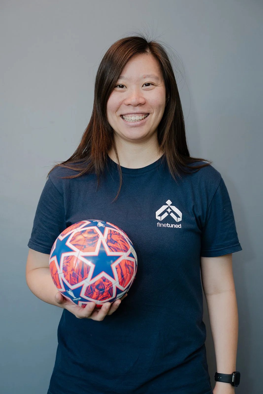 A woman with shoulder-length brown hair smiling at the camera, wearing a navy blue t-shirt with a 'finetuned' logo, holding a colorful soccer ball.