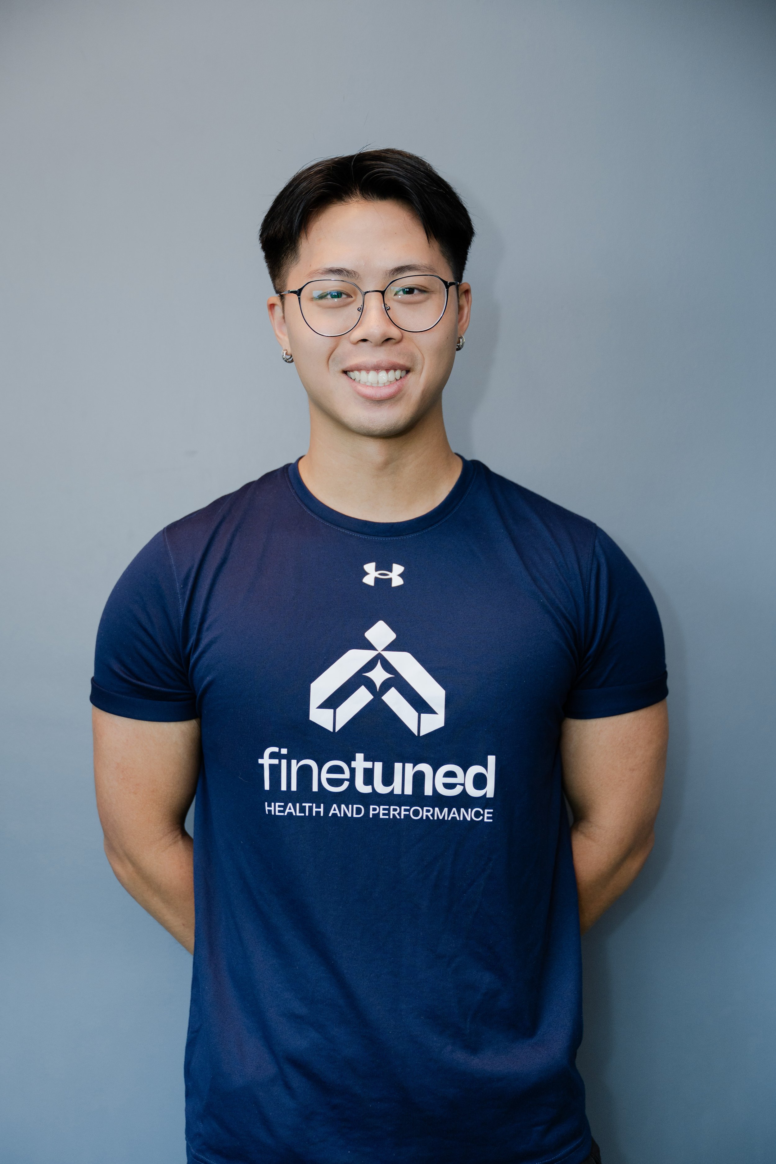 A smiling young man with short black hair, glasses, earrings, wearing a navy blue 'finetuned' T-shirt, standing against a plain gray background.