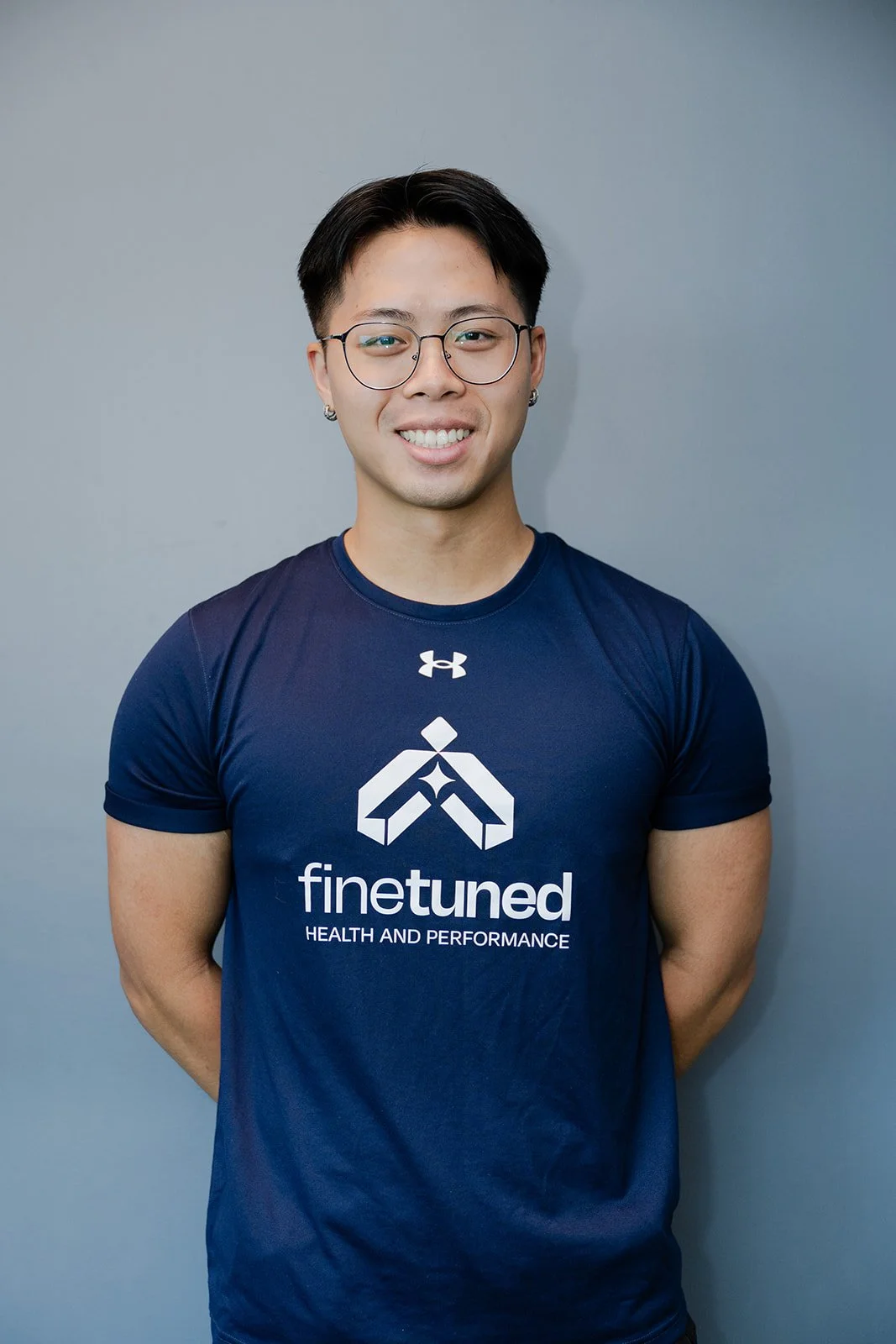 A young man with short dark hair, glasses, and earrings, wearing a navy blue T-shirt with a logo and the words "finetuned HEALTH AND PERFORMANCE," standing against a plain gray wall.