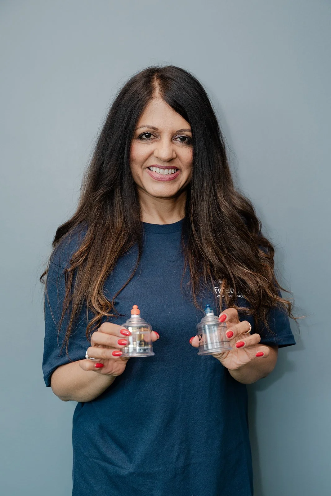 A woman with long dark hair and a blue shirt smiling while holding two baby bottles.