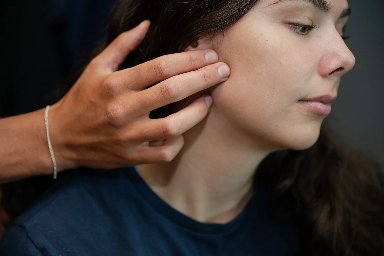 Close-up of a person touching their neck with their hand, showing part of their face, chin, lips, and shoulder, against a neutral background.