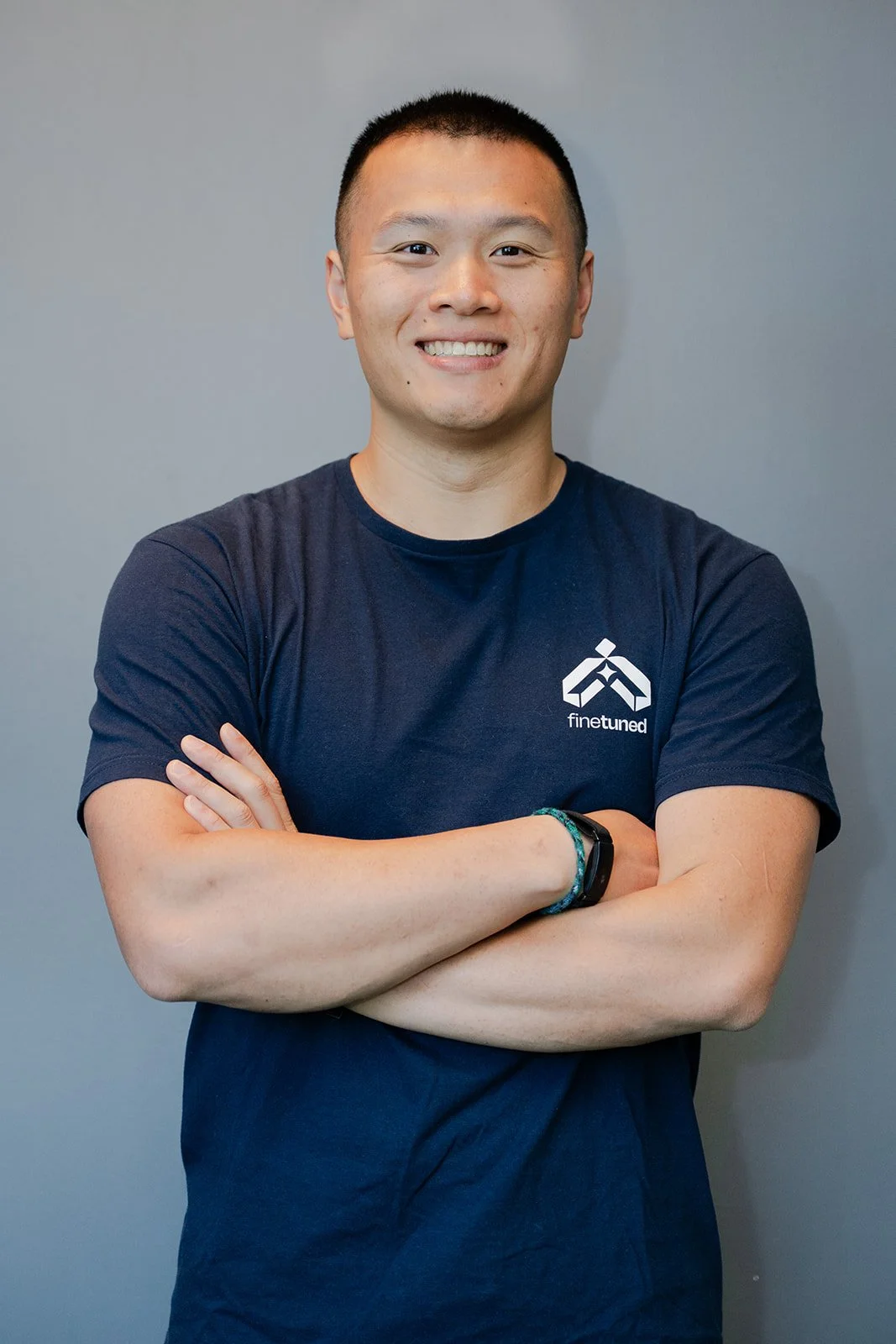 Portrait of a young man with short black hair smiling, wearing a navy blue t-shirt with a white logo and the word 'finetuned' on it, against a neutral gray background.