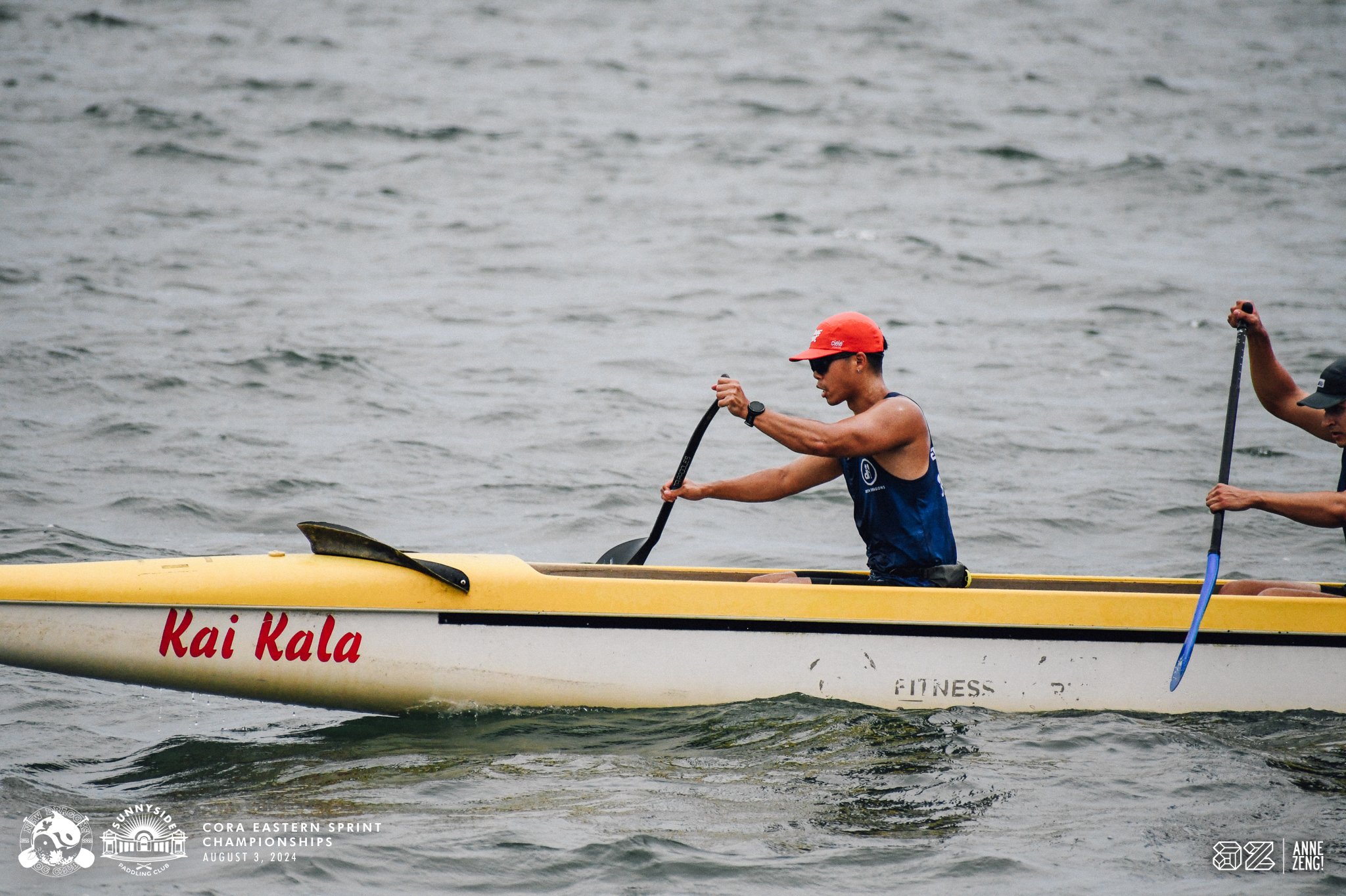 Two people paddling a yellow kayak on a body of water, with the person in the foreground wearing a red cap and sunglasses.