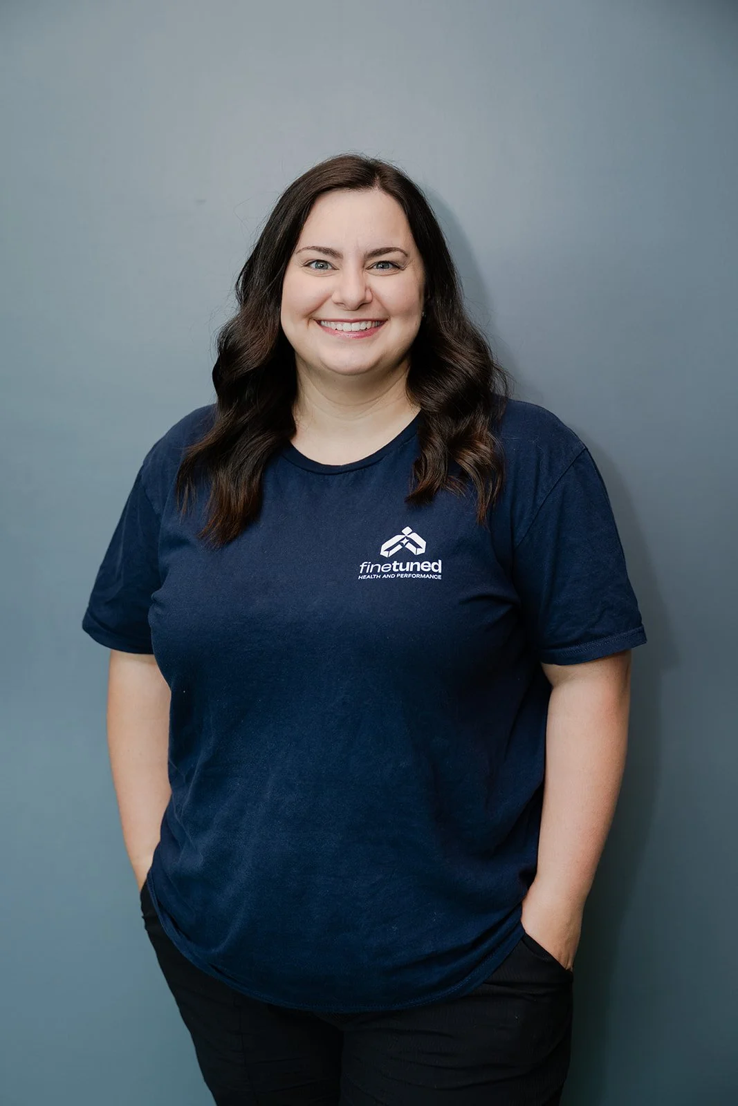 Close-up of a smiling woman with long brown hair and blue eyes wearing a navy blue shirt with a white logo and text that reads 'finetuned' against a plain gray background.