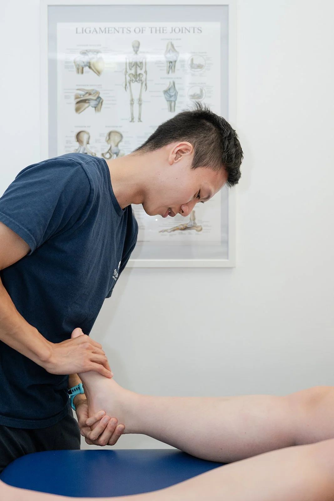 A female chiropractor performing an assessment on a person's ankles, which are lying on a therapy table. There is a model of a human spine in the background.